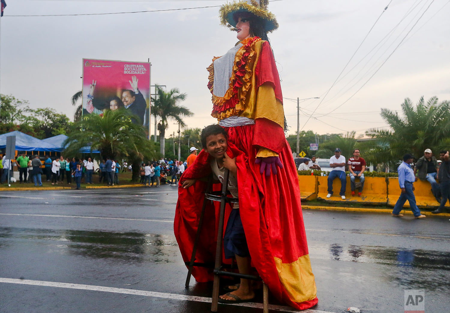  In this May 22, 2018 photo, a youth peers from under a large doll coined "La Gigantona," or The Gigantic, during a pro-government event in Managua, Nicaragua, where a billboard stands behind of President Daniel Ortega and his wife, Vice President Ro