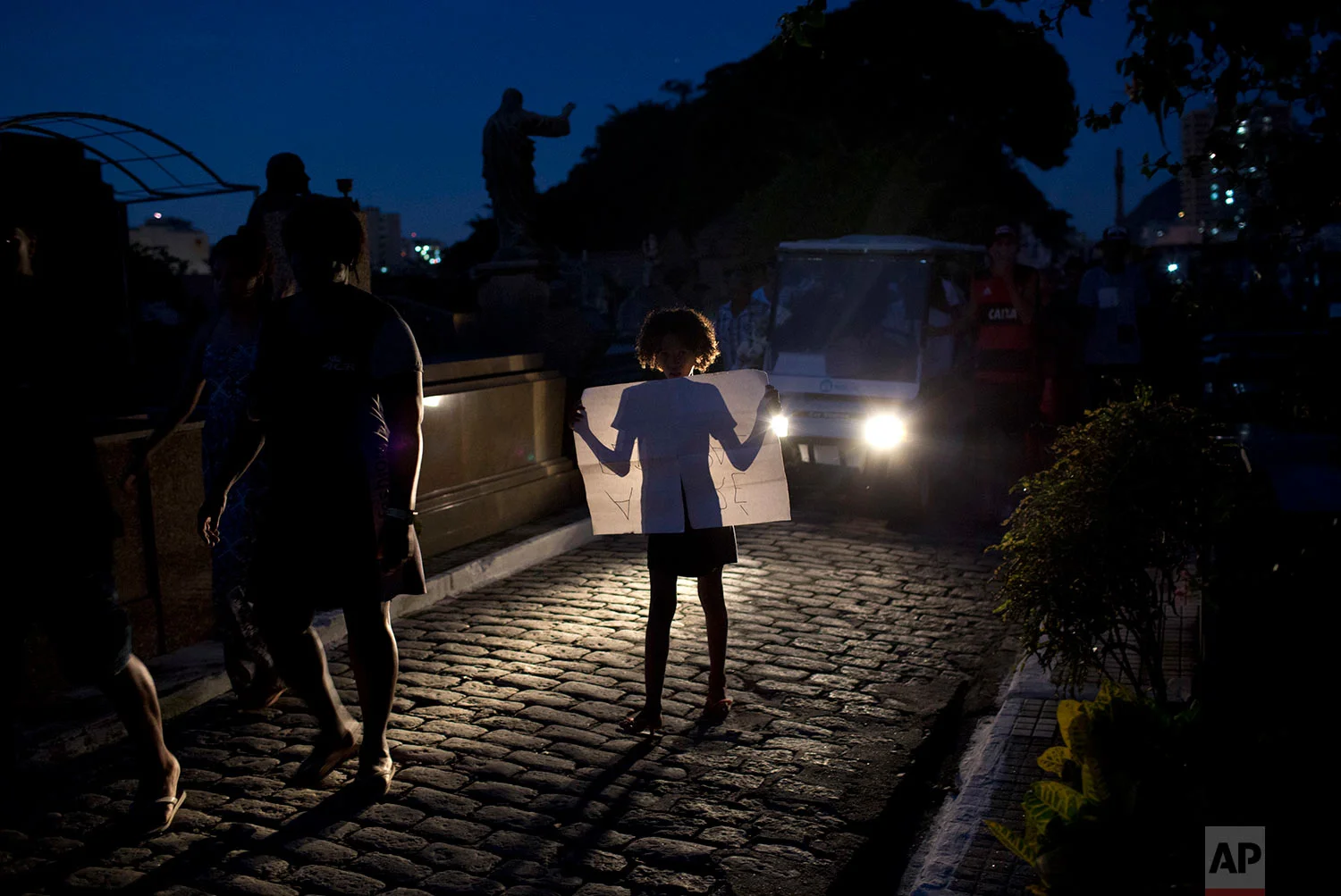 A child holds a sign protesting the death of teenager Marcos Vinicius da Silva, illuminated by the headlights of the vehicle transporting the coffin containing his remains to a cemetery in Rio de Janeiro, Brazil, June 21, 2018. Da Silva is one of two 14-year-olds killed by stray bullets in Rio on Wednesday. (AP Photo/Silvia Izquierdo)