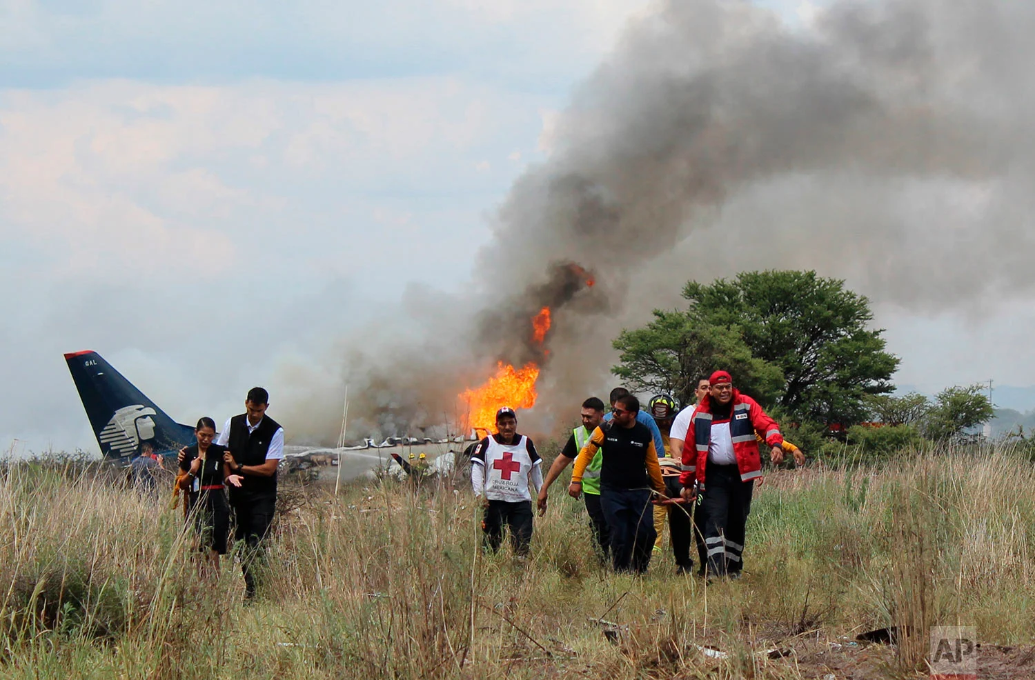 In this photo released by Red Cross Durango communications office, Red Cross workers and rescue workers carry an injured person on a stretcher, right, as airline workers, left, walk away from the site where an Aeromexico airliner crashed in a field near the airport in Durango, Mexico, July 31, 2018. The jetliner crashed while taking off during a severe storm, smacking down in a field nearly intact then catching fire, and officials said it appeared everyone on board escaped the flames. (Red Cross Durango via AP)