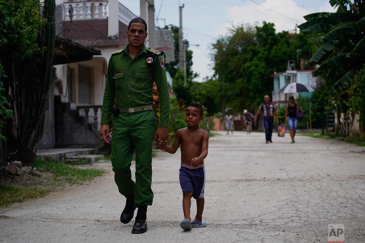 In this July 25, 2018 photo published in August, a Cuban special brigade first lieutenant police officer walks with his son to a neighborhood party where they will cook a large stew, in Guantanamo, Cuba, near the U.S. naval base. It is tradition in Cuba for neighbors to pool their efforts to cook a large stew, using pork. (AP Photo/Ramon Espinosa)