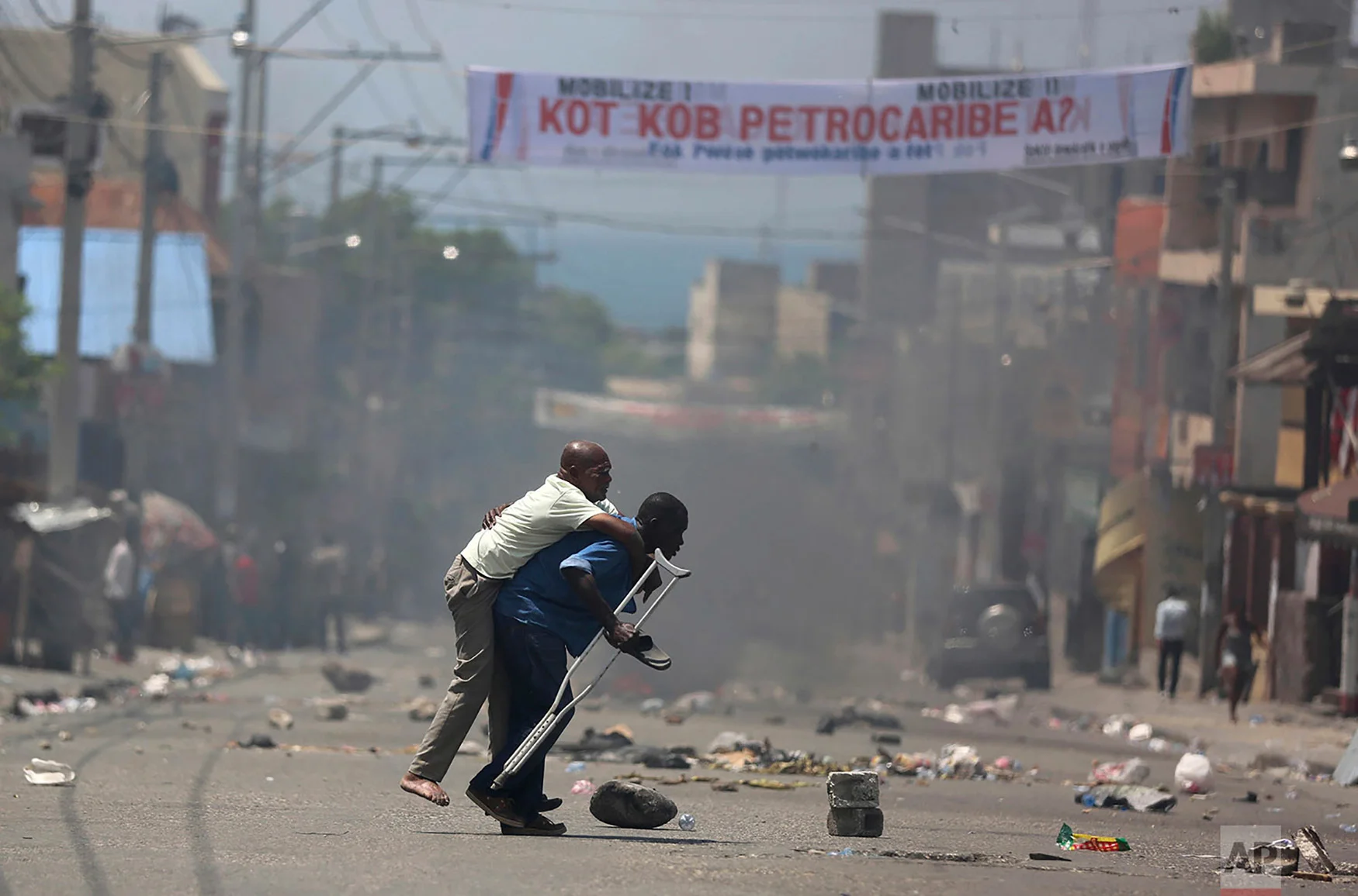 A handicapped man is carried across the street amid police firing tear gas at protesters who are demanding to know how PetroCaribe funds have been used by the current and past administrations, in Port-au-Prince, Haiti, Sept. 9, 2018. Much of the financial support to help Haiti rebuild after the 2010 earthquake comes from Venezuela's PetroCaribe fund, a 2005 pact that gives suppliers below-market financing for oil and is under the control of the central government. (AP Photo/Dieu Nalio Chery)