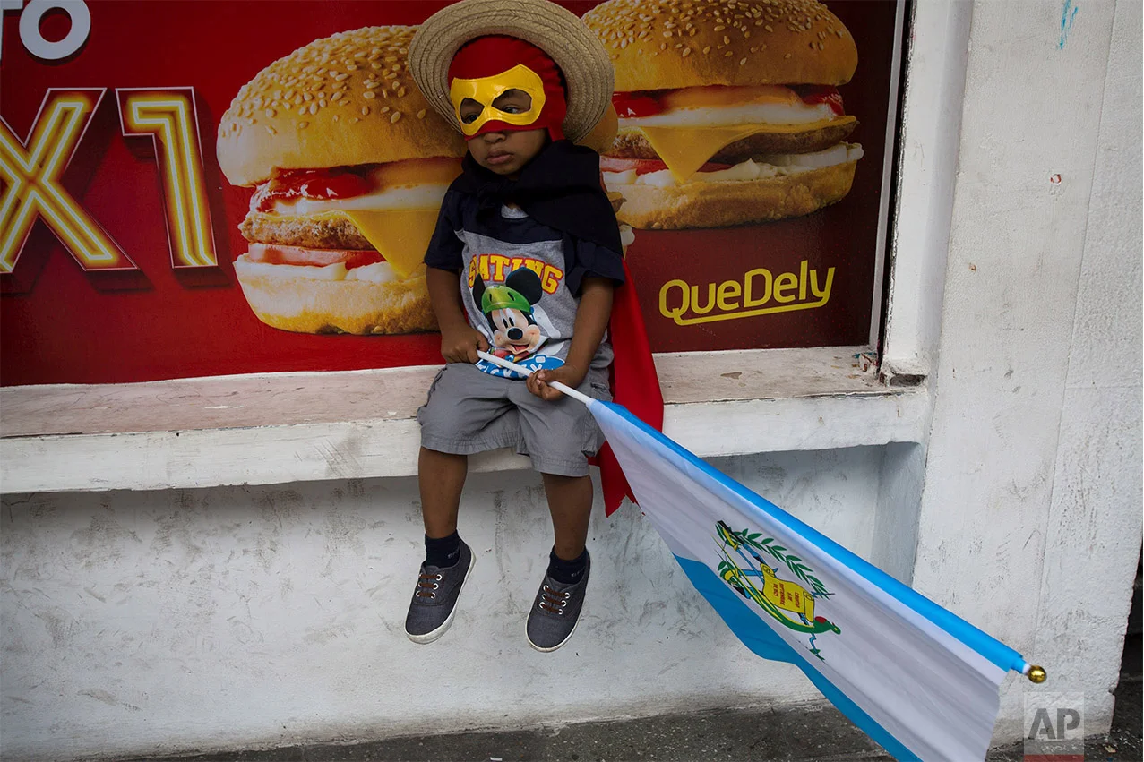 A boy in costume clutching a Guatemalan flag sits outside a local fast food restaurant as he waits with his father for a march to start against Guatemalan President Jimmy Morales and corruption in Guatemala City, Sept. 20, 2018. Thousands marched to protest Morales' decision to end the work of a U.N. anti-corruption commission that has helped lead high-profile graft probes targeting dozens of powerful people, including one involving Morales. (AP Photo/Moises Castillo)