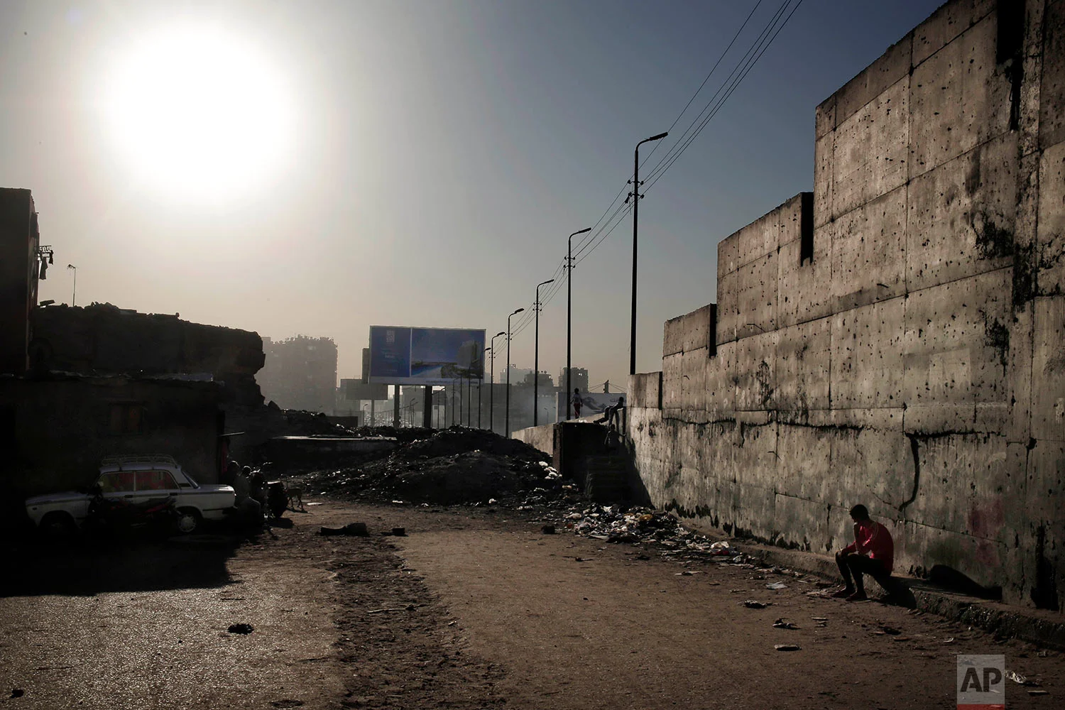 In this Oct. 28, 2018 photo, a man sits under a wall that separates the ring road from slum area Ezbet Khairallah, in Cairo, Egypt. (AP Photo/Nariman El-Mofty)