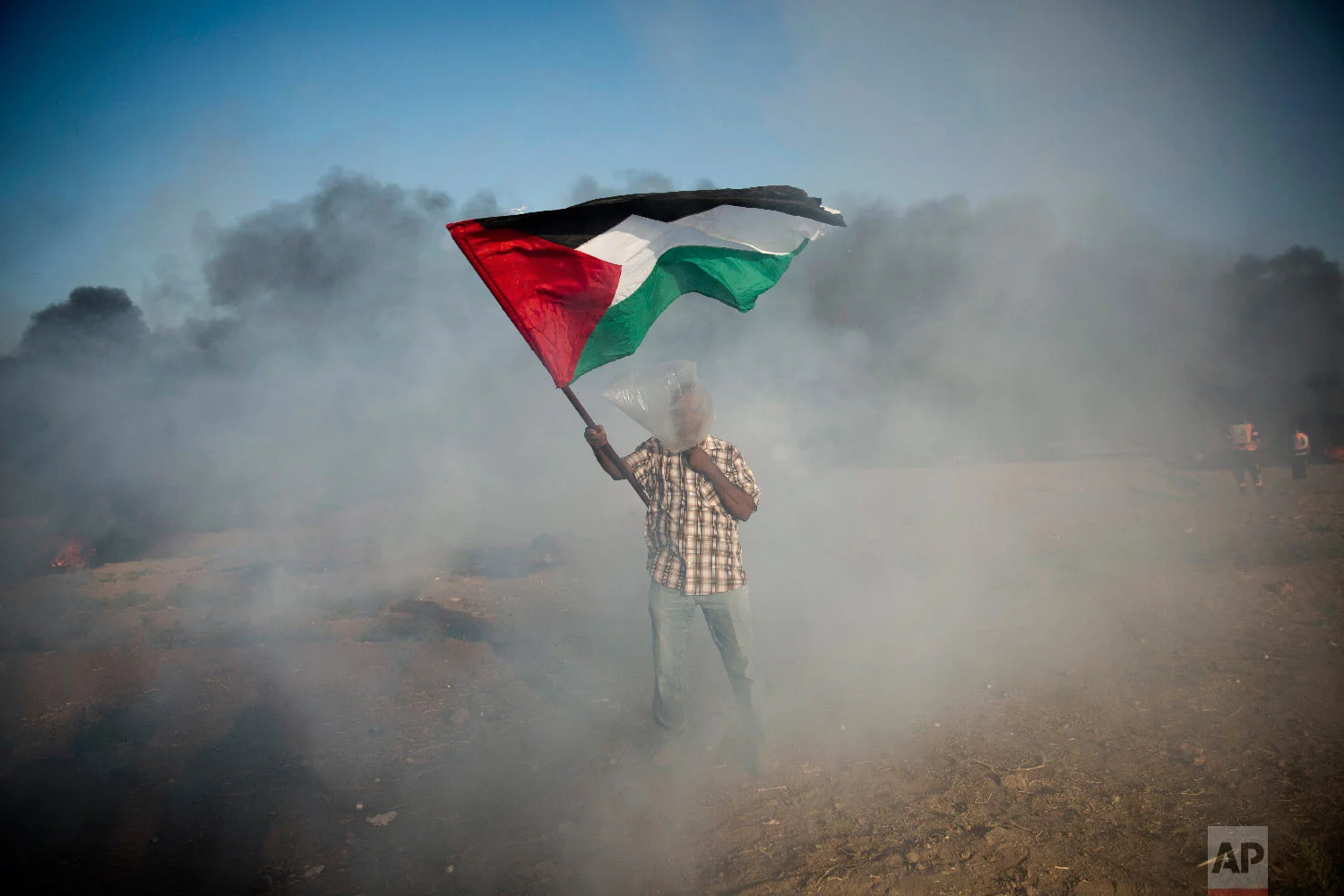A Palestinian protester wears a plastic bag on his head as a protection from teargas, waves a national flag during a protest at the Gaza Strip's border with Israel, Aug.10, 2018. (AP Photo/Khalil Hamra)