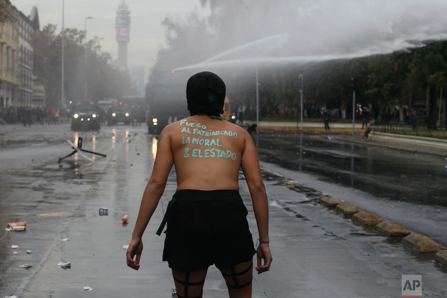 In this May 16, 2018 photo, a topless woman with the Spanish message on her back: "Fire to the patriarchy, morals and the state" confronts police spraying a water cannon in Santiago, Chile. Students protested sexual abuse, sexism and demanded equal opportunities for woman in education. (AP Photo/Luis Hidalgo)