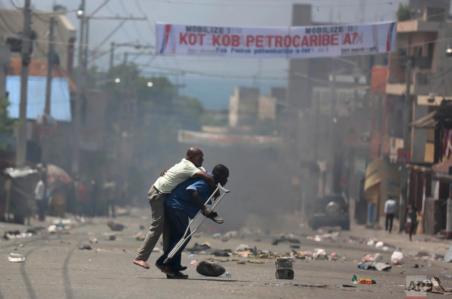 In this Sept. 9, 2018 photo, a handicapped man on crutches is carried amid police tear gas as protesters demand to know how the government spent Petro Caribe funds, intended to help Haiti rebuild after the 2010 earthquake, in Port-au-Prince, Haiti. Protesters say projects are unfinished and officials inflated costs to steal money. (AP Photo/Dieu Nalio Chery)