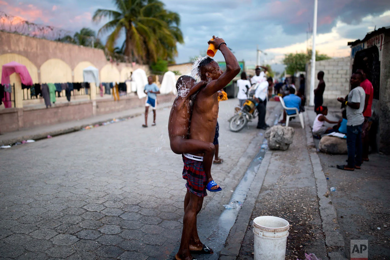 In this Nov. 13, 2018 photo, a man takes a public bucket shower with his nephew on his back in the Cite Soleil slum of Port-au-Prince, Haiti. Most residents in Cite Soleil do not have bathrooms, and bathe outdoors. (AP Photo/Dieu Nalio Chery)