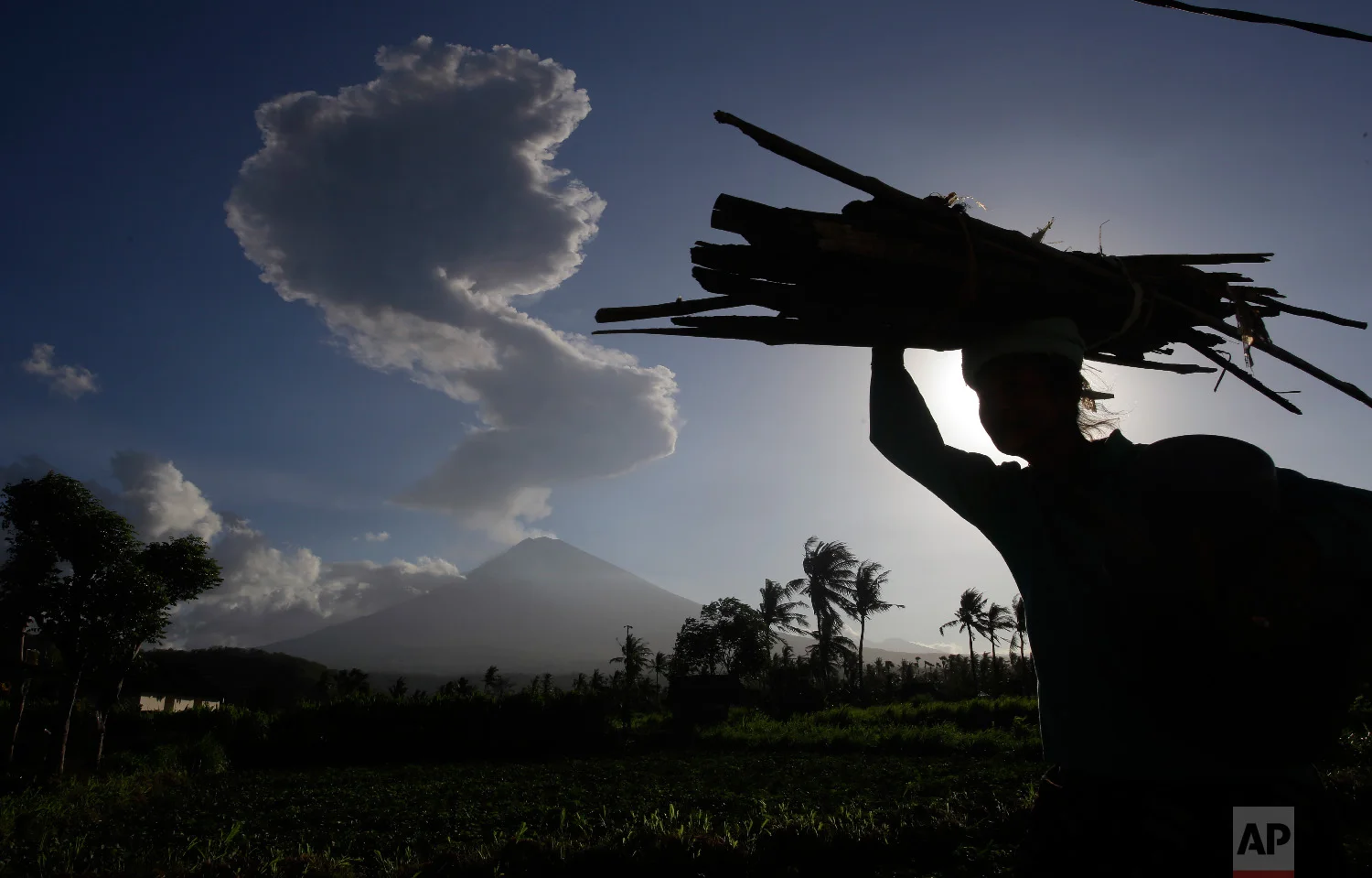 A woman carries wood past as the Mount Agung spews ash and smoke in the background in Karangasem, Bali, Indonesia on July 5, 2018. (AP Photo/Firdia Lisnawati)