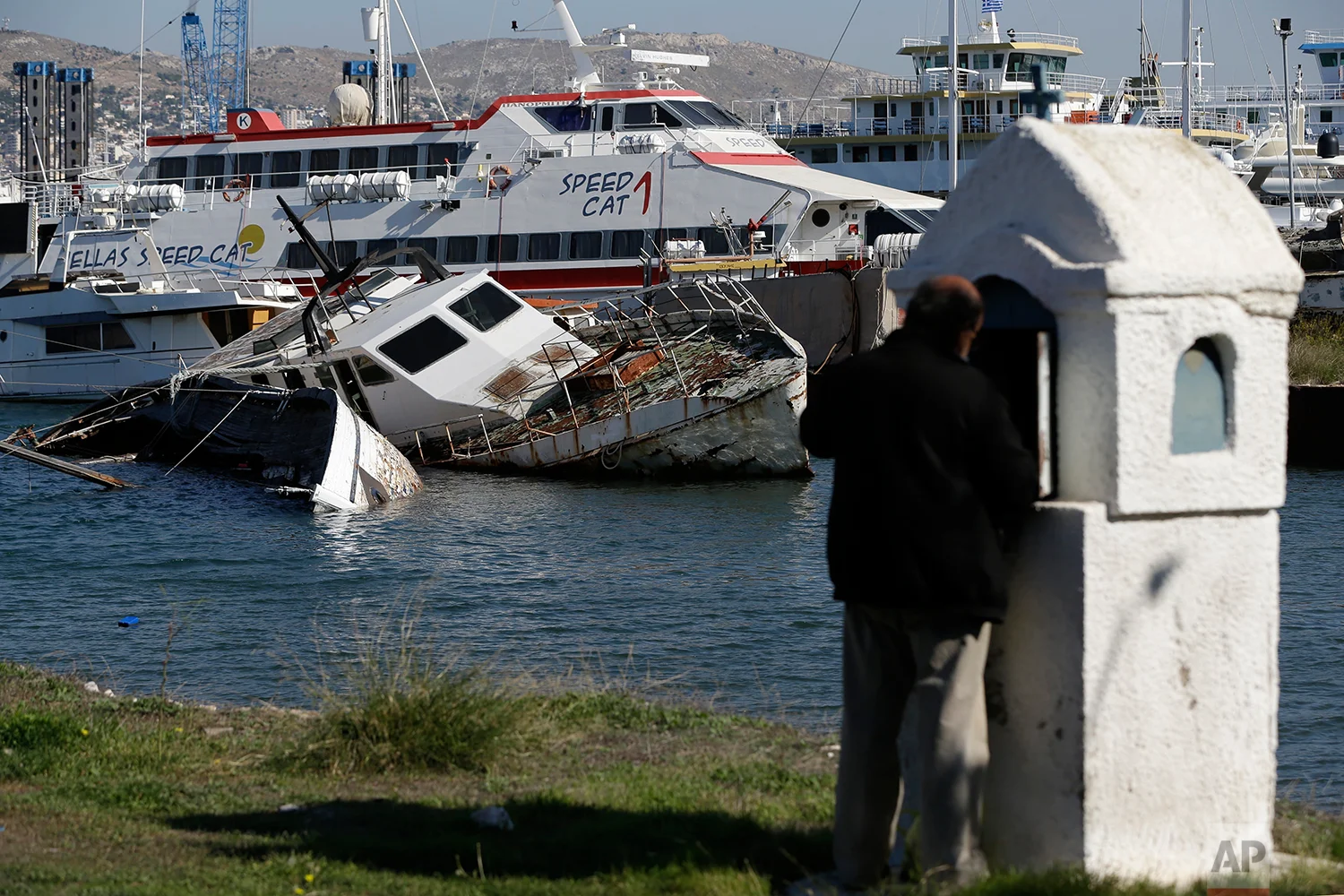 In this Tuesday, Nov. 6, 2018 photo, a half sunken small boat is photographed near a shipyard in Salamina island, west of Athens as a man lights a candle at a roadside shrine. (AP Photo/Thanassis Stavrakis)