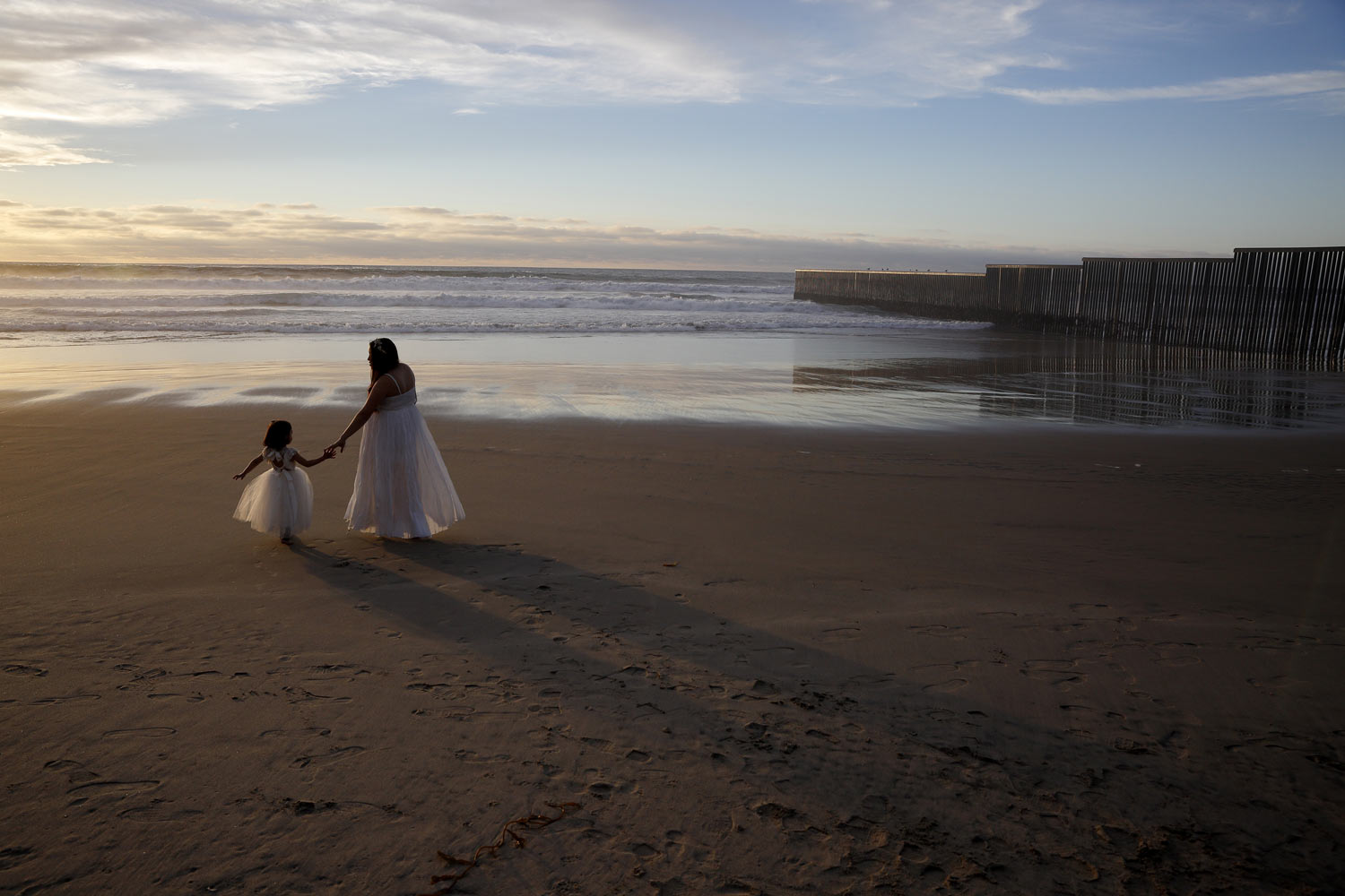 Ileze Dariel, of Tijuana, Mexico, reaches for the hand of her daughter, Jimena, as they wait for their photographer while taking family pictures on the beach next to the border wall, right, Wednesday, Jan. 9, 2019, in Tijuana, Mexico. (AP Photo/Gregory Bull)