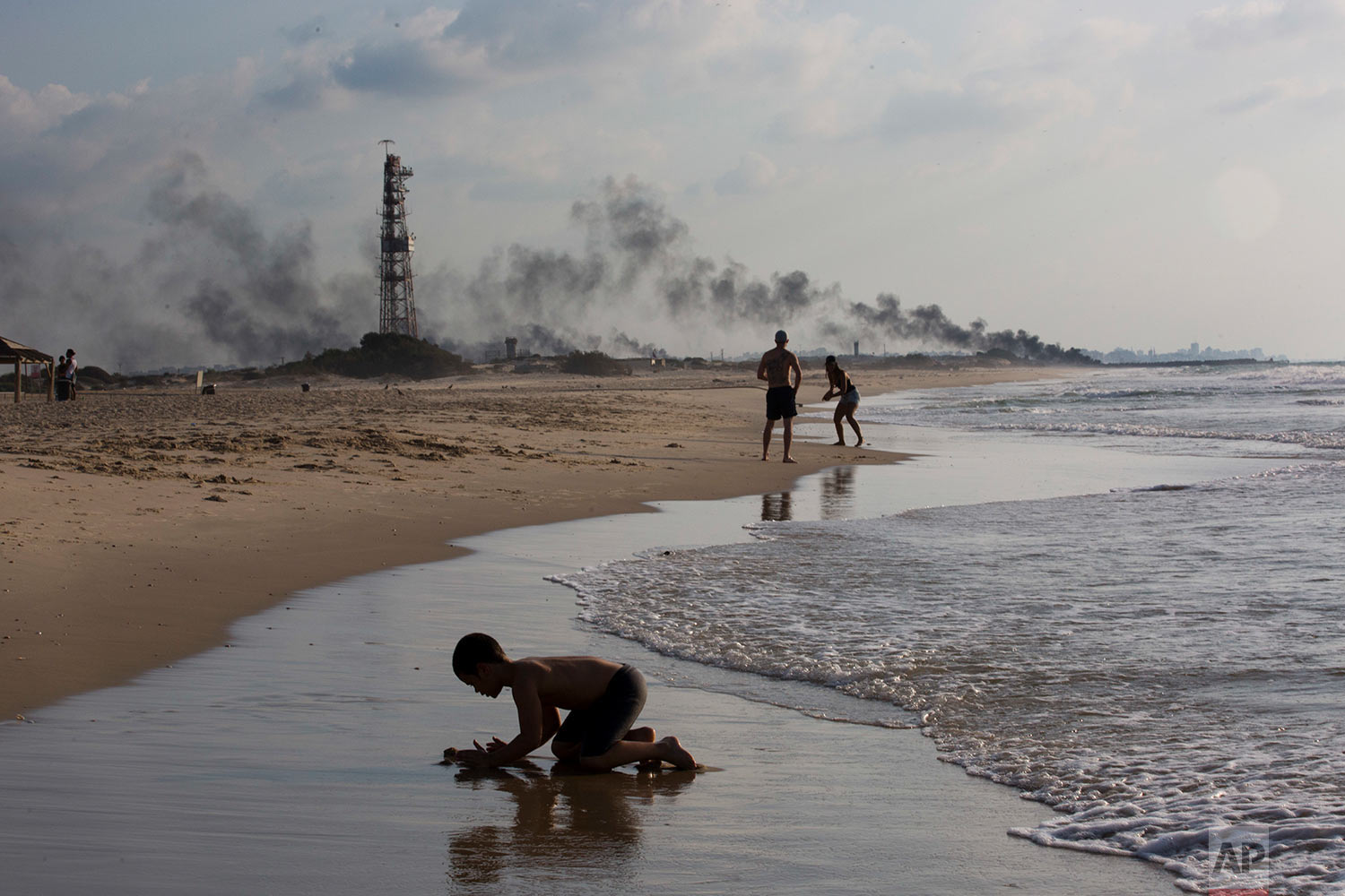 Israelis enjoy the day in Zikim beach, near kibbutz Zikim, on the Israel and Gaza border, as in the background black smoke rises from the tires set on fire by Palestinian protesters near Beit Lahiya, northern Gaza Strip, Oct. 8, 2018. (AP Photo/Oded Balilty)