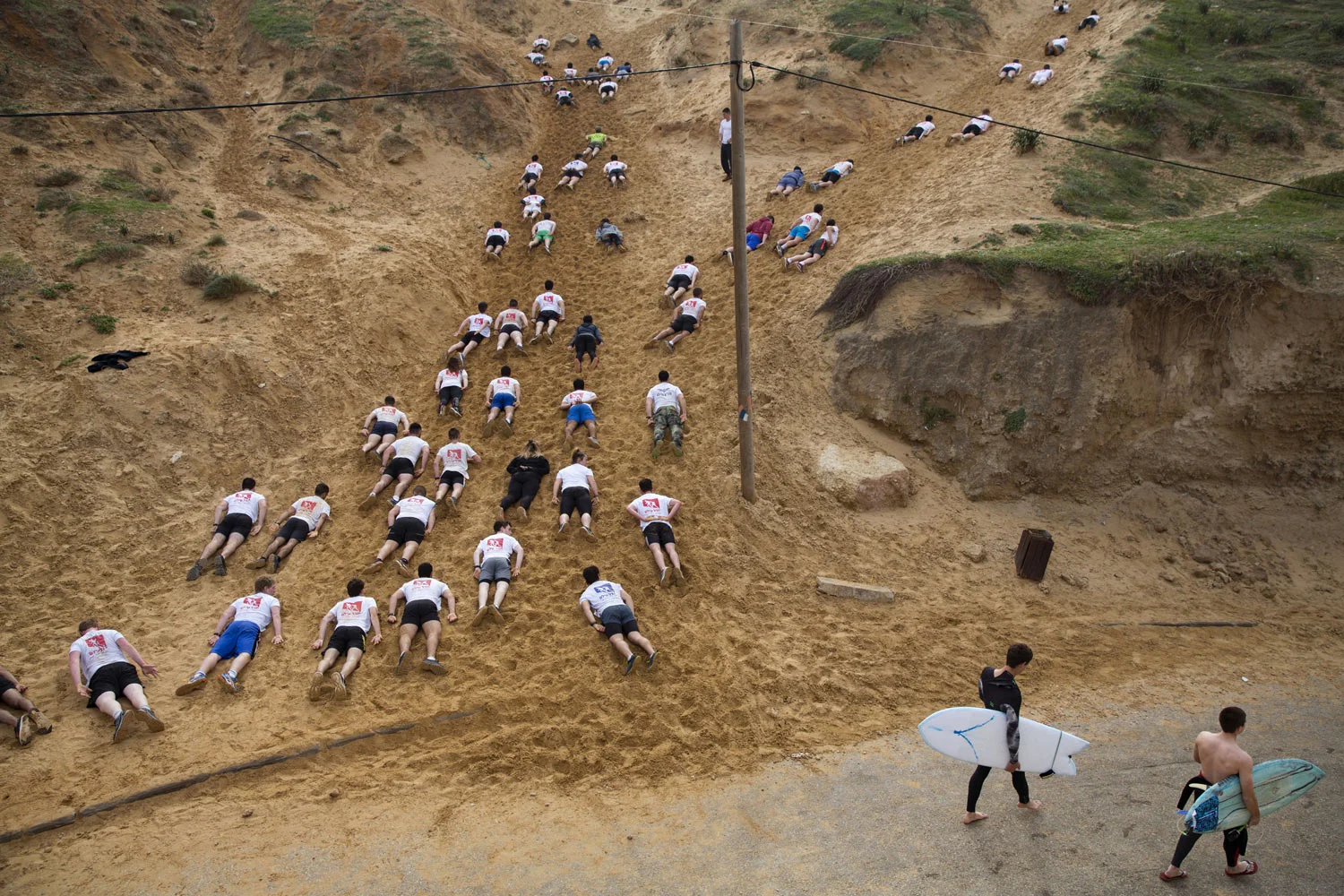 Two surfers walk towards the Mediterranean Sea as high school seniors preparing to join the Israeli military later in the year crawl along sand dunes during an exercise at a privately run training camp in Herzliya, Israel, Friday, Feb. 1, 2019. (AP