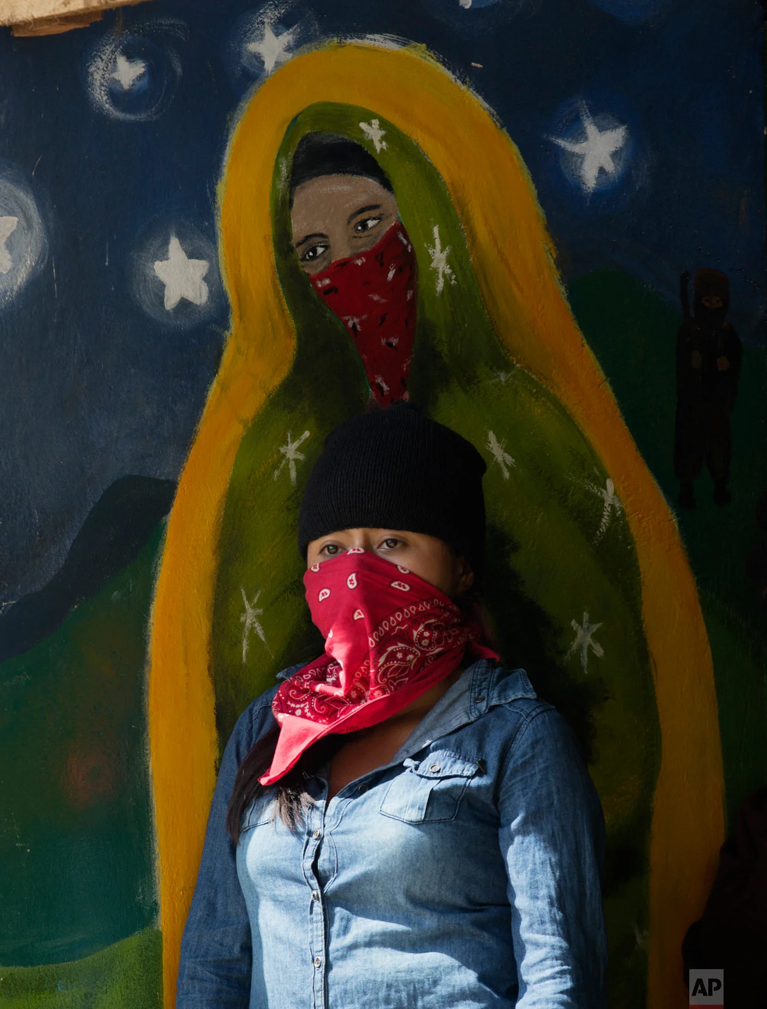 A masked, female member of the Zapatista National Liberation Army (EZLN) stands by a mural depicting a masked version of the Virgin of Guadalupe, at an event marking the 25th anniversary of the Zapatista uprising in La Realidad, Chiapas, Mexico, Jan. 1, 2019. (AP Photo/Eduardo Verdugo)