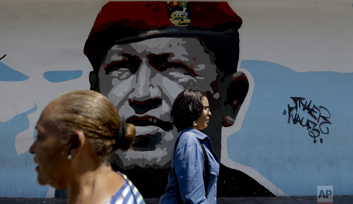 Pedestrians walk past a mural featuring Venezuela's late President Hugo Chavez, in Caracas, Venezuela, Tuesday, March 19, 2019. Chavez, a former military officer, declared a socialist "revolution" after coming to power in 1999. He was succeeded after his 2013 death by his protege, Nicolas Maduro. (AP Photo/Natacha Pisarenko)