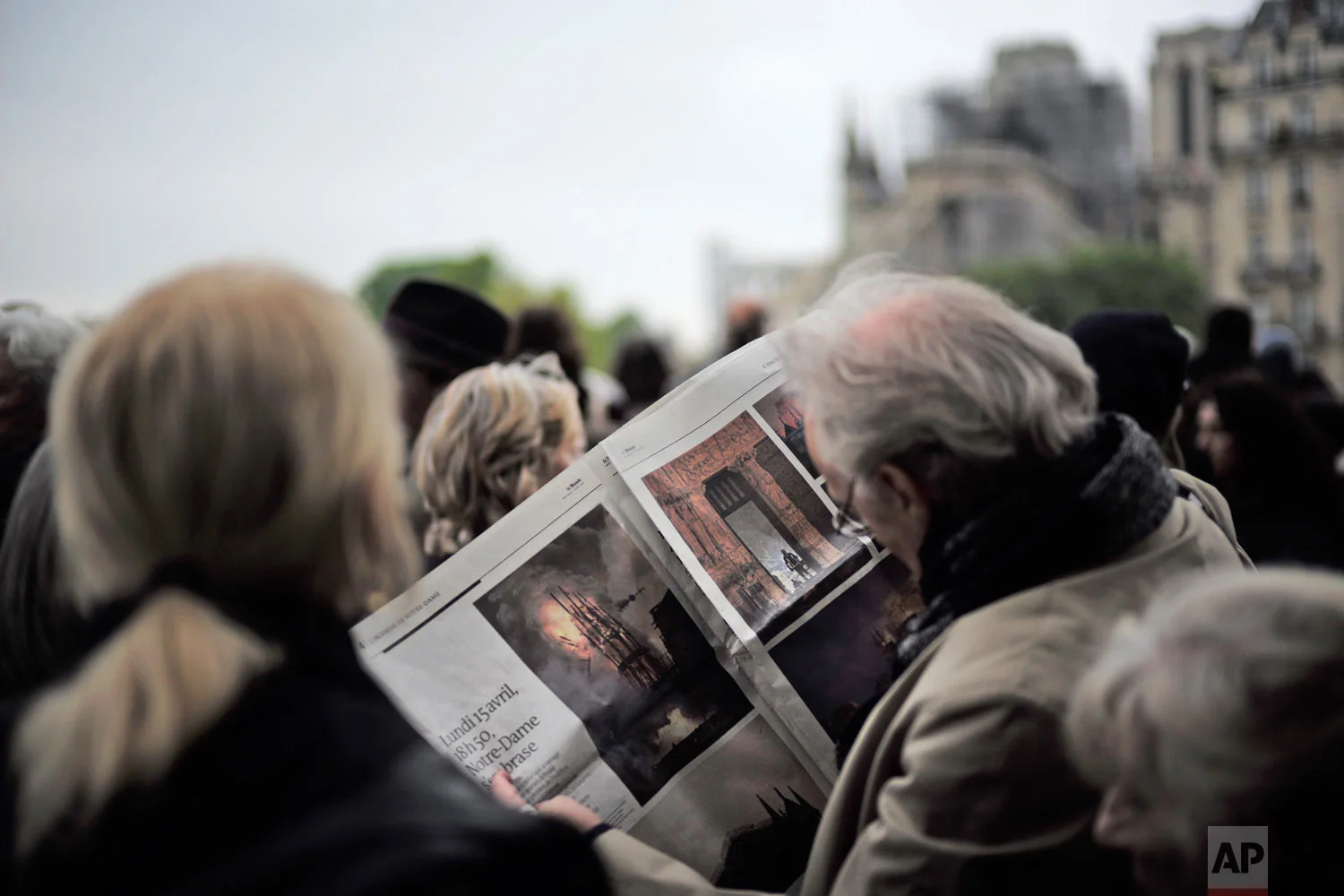 A man looks at pictures of the burning Notre Dame Cathedral in a newspaper as he and others gather near the damaged cathedral, rear, after the fire in Paris, April 16, 2019. (AP Photo/Kamil Zihnioglu)