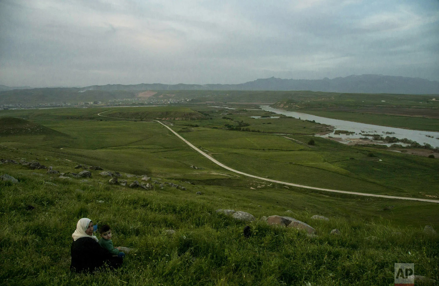 A grandmother sits in the grass with her grandson on a hill overlooking the Euphrates River as families picnic on May Day, Wednesday, May 1, 2019, in Ein Diwar, Derik, Syria. (AP Photo/Baderkhan Ahmad)