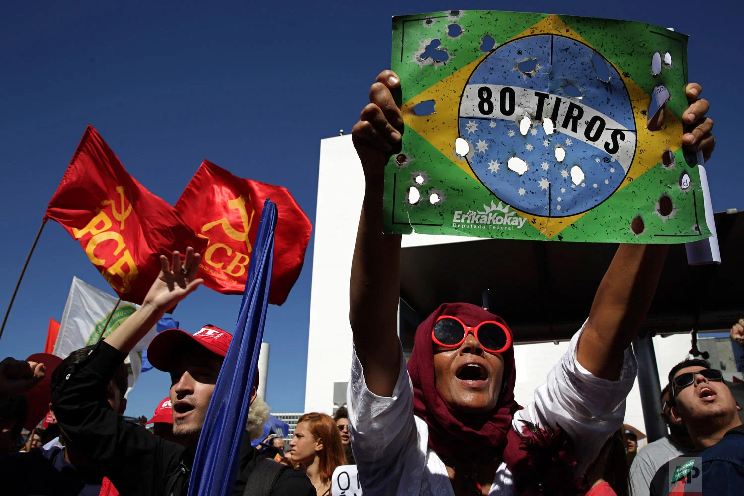 A demonstrator holds up a poster of a Brazilian flag marred with bullet holes and a message that reads in Portuguese: "80 shots," in reference to soldiers who last month fired 80 shots at a car carrying a family, killing the driver, during a protest against education budget cuts in Brasilia, Brazil, Thursday, May 30, 2019. (AP Photo/Eraldo Peres)