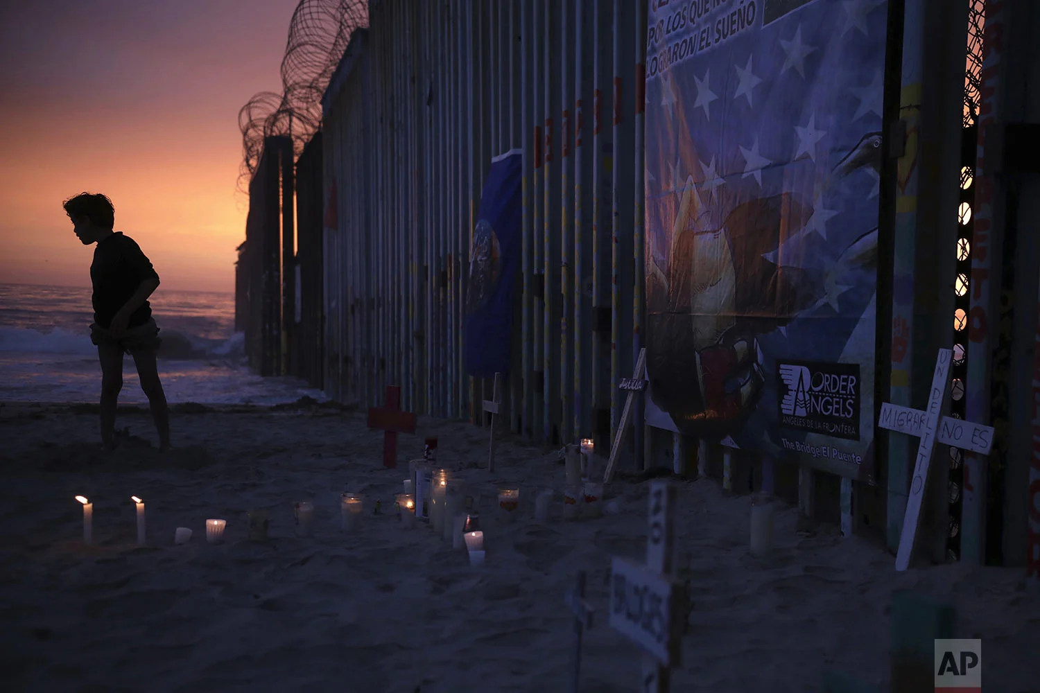A youth stands by the border fence that separates Mexico from the United States, near a makeshift memorial for migrants who have died during their journey toward the U.S., in Tijuana, Mexico, late Saturday, June 29, 2019. On the border fence at right hangs a cartoon depiction of a news photograph of the bodies of Salvadoran migrant Óscar Alberto Martínez Ramírez and his daughter Valeria, photographed on the banks of the Rio Grande between Matamoros, Mexico, and Brownsville, Texas, after they drowned on Sunday, June 23. (AP Photo/Emilio Espejel)