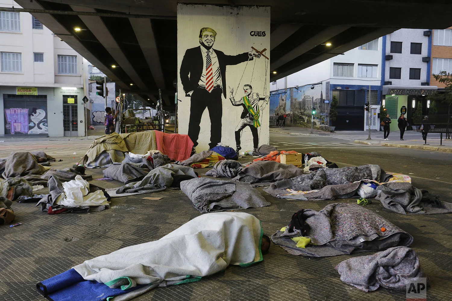 Homeless people sleep under a bridge in front of a mural depicting U.S. President Donald Trump as a puppeteer manipulating Brazil's President Jair Bolsonaro, in downtown Sao Paulo, Brazil, Tuesday, July 23, 2019. Bolsonaro said last week that severe hunger is not a problem for the country. He acknowledged that things could "go bad" and people might "not eat well." But, he added: "To speak of starving in Brazil is a big lie." (AP Photo/Nelson Antoine)