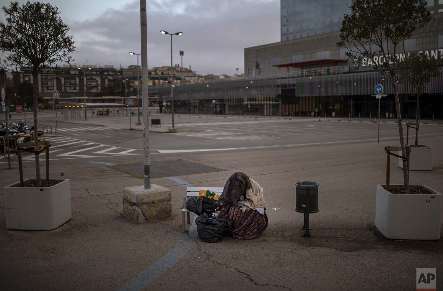  In this Sunday, March 22, 2020, a man of sub-Saharan Africa covers himself with clothes and blankets as he sits in a bench of an empty parking outside the train station in Barcelona, Spain. (AP Photo/Emilio Morenatti) 