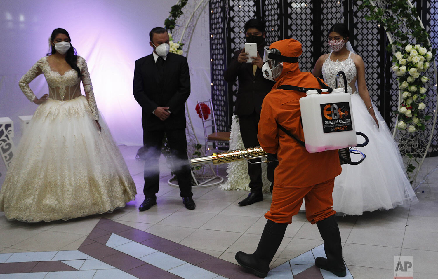  A man wearing a full protection suit sprays disinfectant on two couples in bridal dresses and suits, inside a ballroom after a mock wedding during the partial lifting of restrictions amid the COVID -19 pandemic in La Paz, Bolivia, Oct. 7, 2020. (AP 