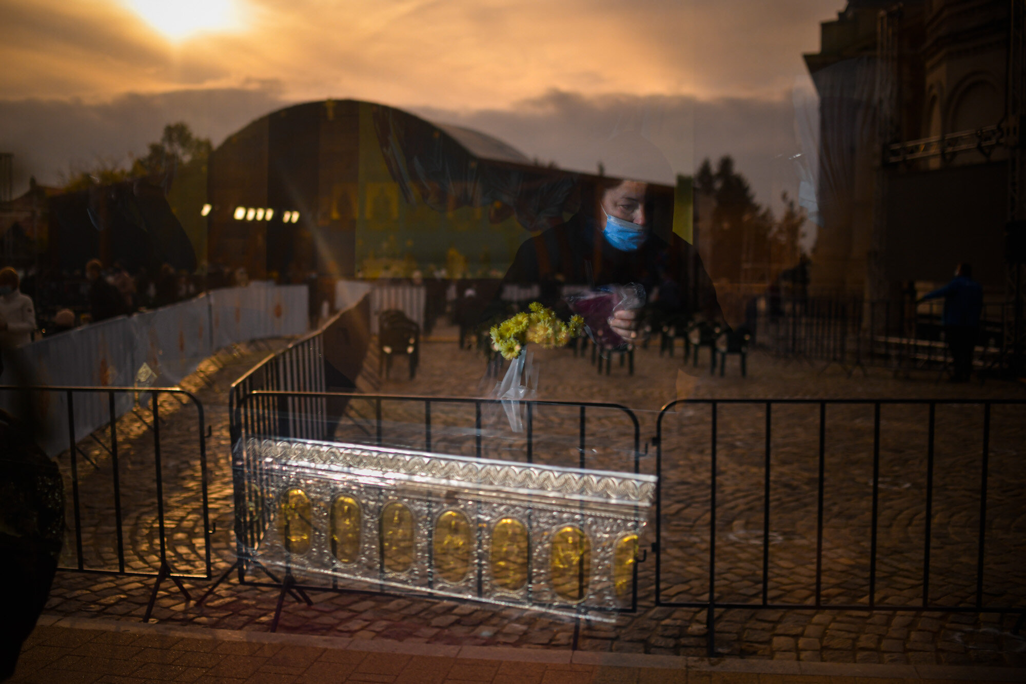 A woman wearing a mask to protect against infection from COVID-19 is reflected in a tinted chapel window, along with a metal casing said to contain the remains of St. Dimitrie of Basarabov, the patron saint of the Romanian capital, in Bucharest, Romania on Oct. 25, 2020. The feast of St. Dimitrie of Basarabov, which usually lasts for a week and draws up to 100,000 people, was cut way back this year due to the coronavirus. (AP Photo/Andreea Alexandru)