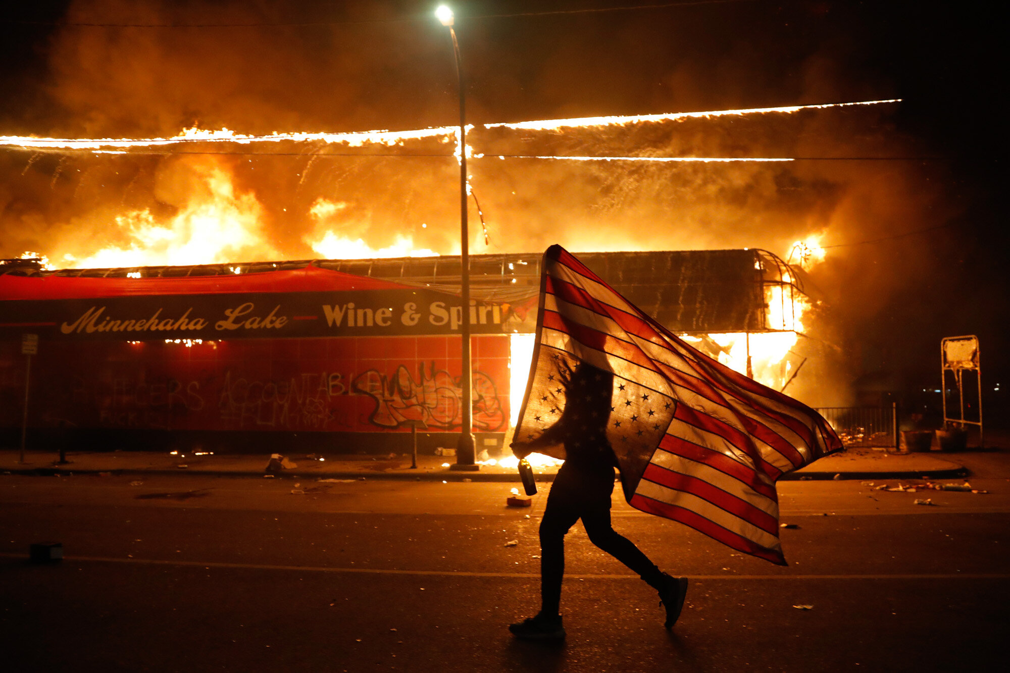 A protester carries a U.S. flag upside down as he walks past a burning building in Minneapolis on May 28, 2020, during a protest over the death of George Floyd, a Black man who died after a white Minneapolis police officer pressed a knee into his neck for several minutes. (AP Photo/Julio Cortez)