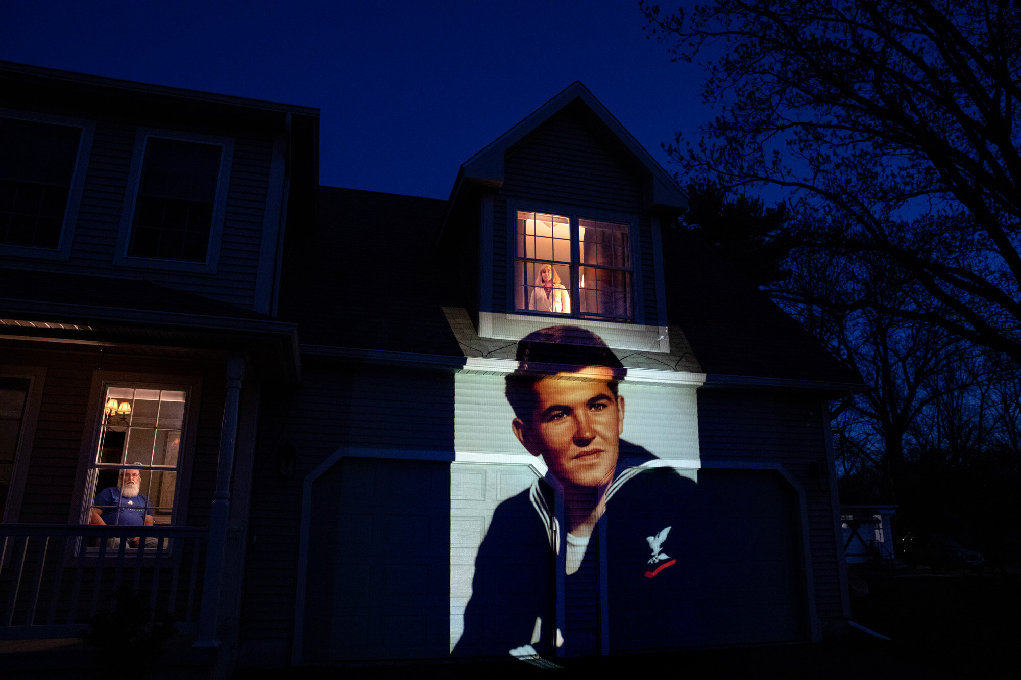 An image of U.S. Navy veteran Stephen Kulig is projected onto the home of his daughter, Elizabeth DeForest, as she looks out the window of a spare bedroom while her husband, Kevin, sits downstairs, in Chicopee, Mass., on May 3, 2020. Kulig, a resident of the Soldier's Home in Holyoke, Mass., died from COVID-19 at the age of 92. (AP Photo/David Goldman)