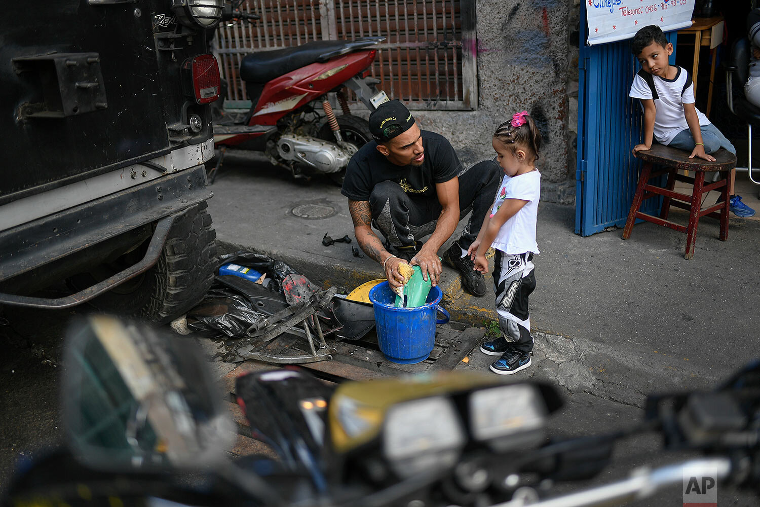  Motorcycle stuntman Pedro Aldana cleans motorcycle parts with his daughter Alanis as his young neighbor and admirer, 6-year-old Milan Sandoval Ramos, watches from a chair outside their home in the Catia neighborhood of Caracas, Venezuela, Thursday, 