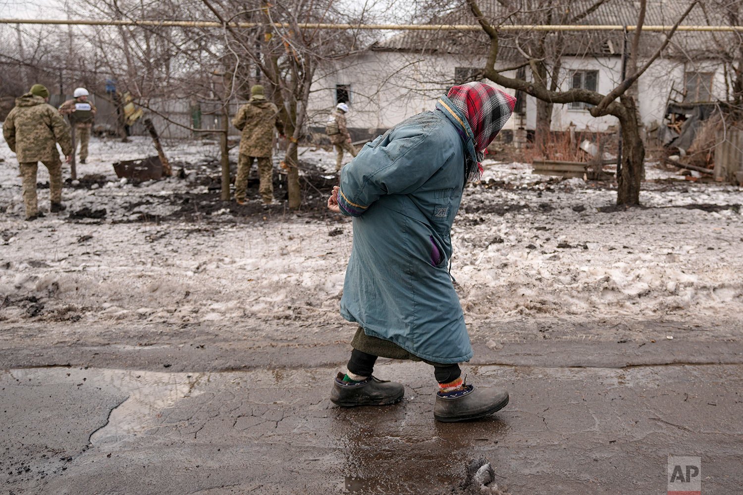  An elderly woman walks by as members of the Joint Centre for Control and Coordination on ceasefire of the demarcation line, or JCCC, survey a crater and damage to a house from artillery shell that landed in Vrubivka, one of the at least eight that h