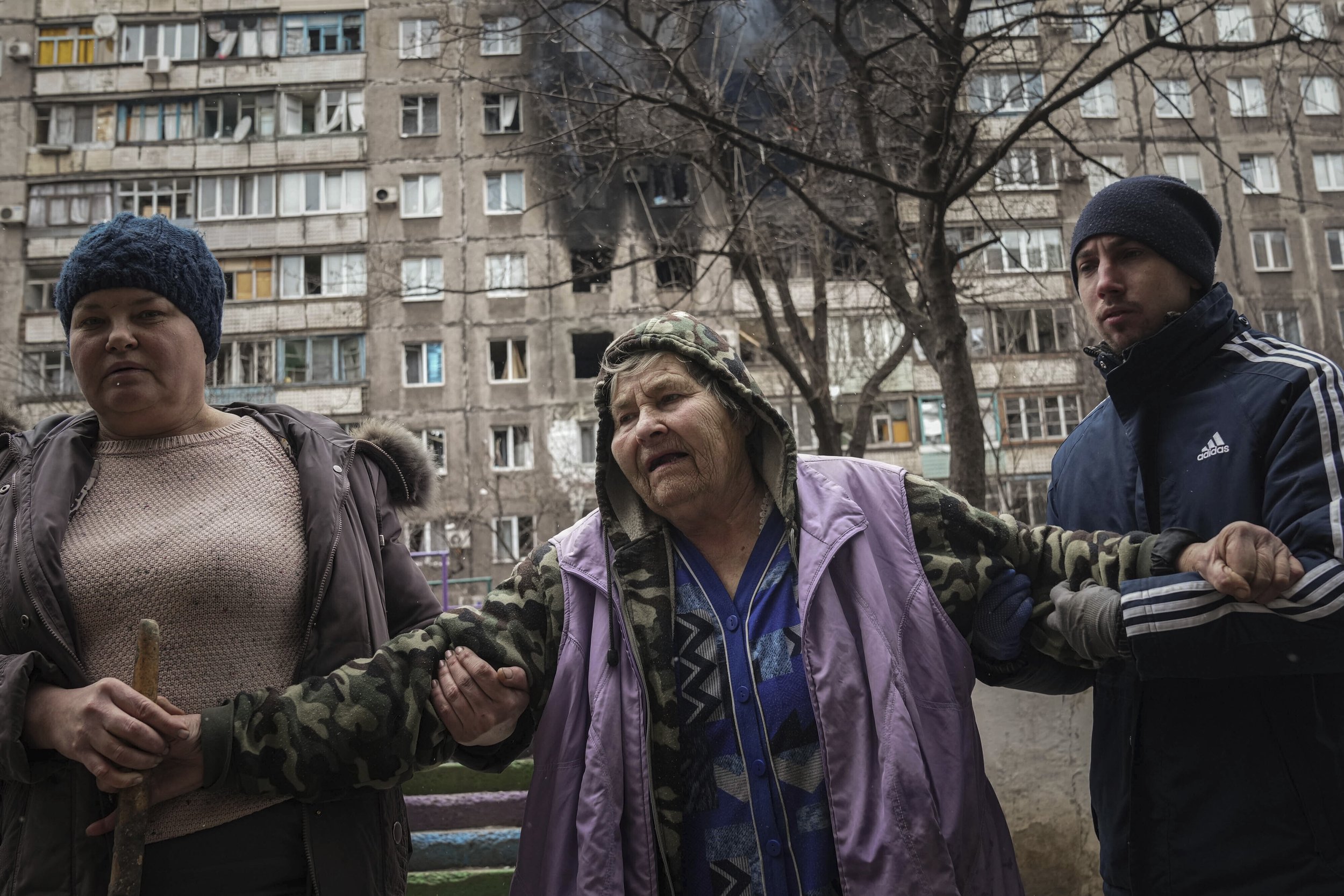  People help an elderly woman to walk in a street with an apartment building hit by shelling in the background in Mariupol, Ukraine, Monday, March 7, 2022. (AP Photo/Evgeniy Maloletka) 
