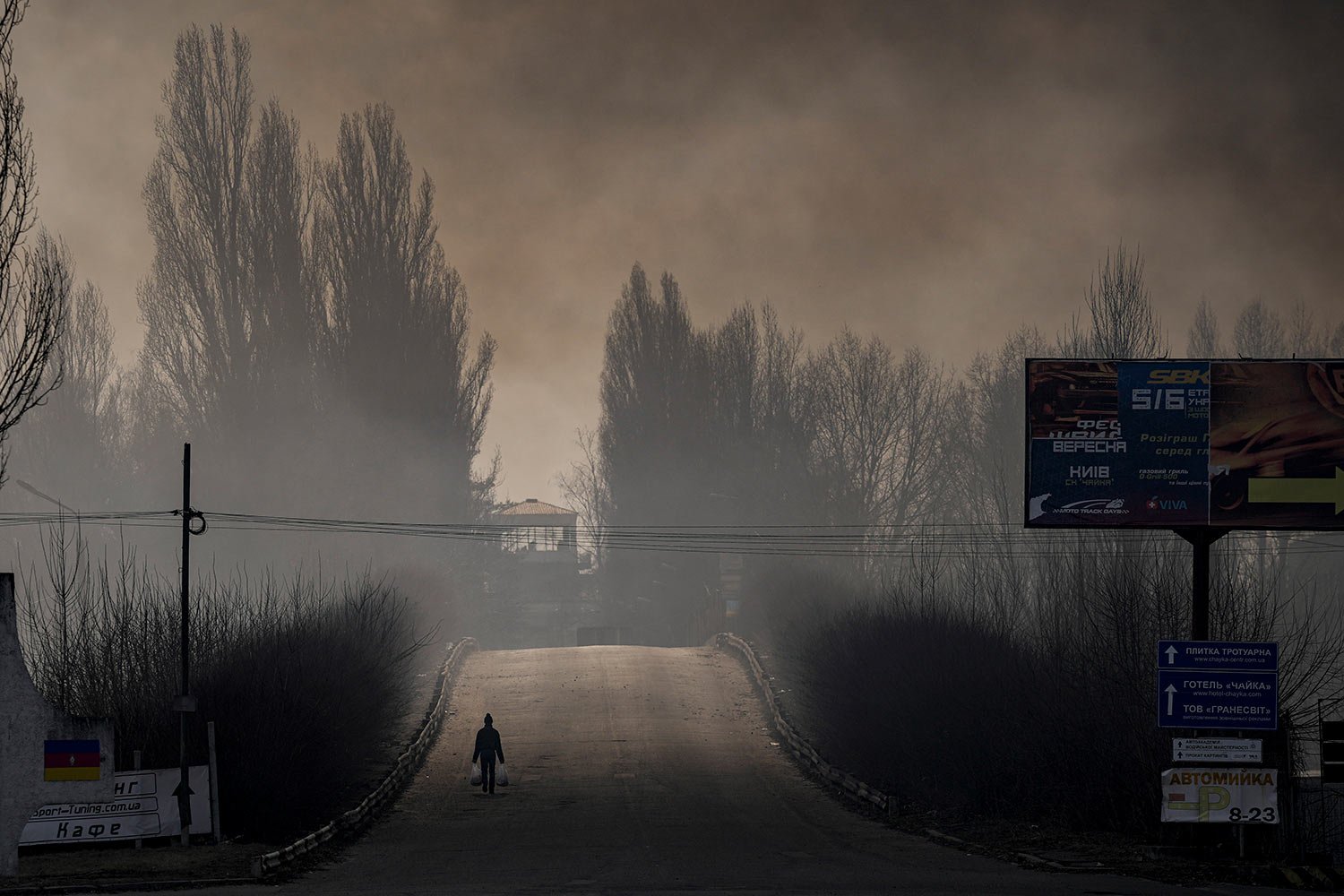  A man carries shopping bags as heavy smoke from a warehouse destroyed by Russian bombardment casts shadows on the road outside Kyiv, Ukraine,Thursday, March 24, 2022. (AP Photo/Vadim Ghirda) 