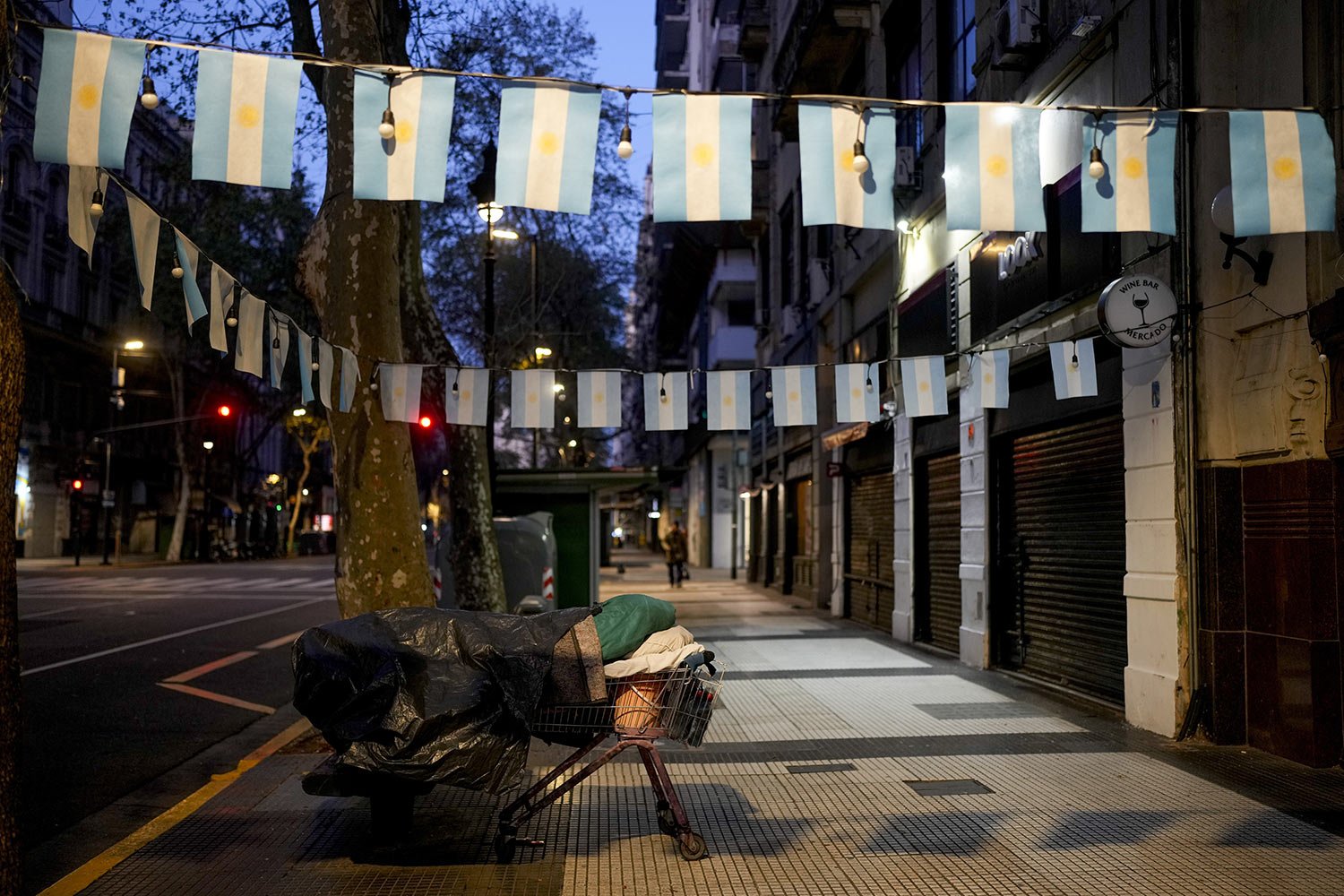 A homeless person sleeps on a bed made of a shopping cart and bench on a sidewalk decorated with Argentine flags in Buenos Aires, Argentina, early Sept. 27, 2023. (AP Photo/Natacha Pisarenko) 