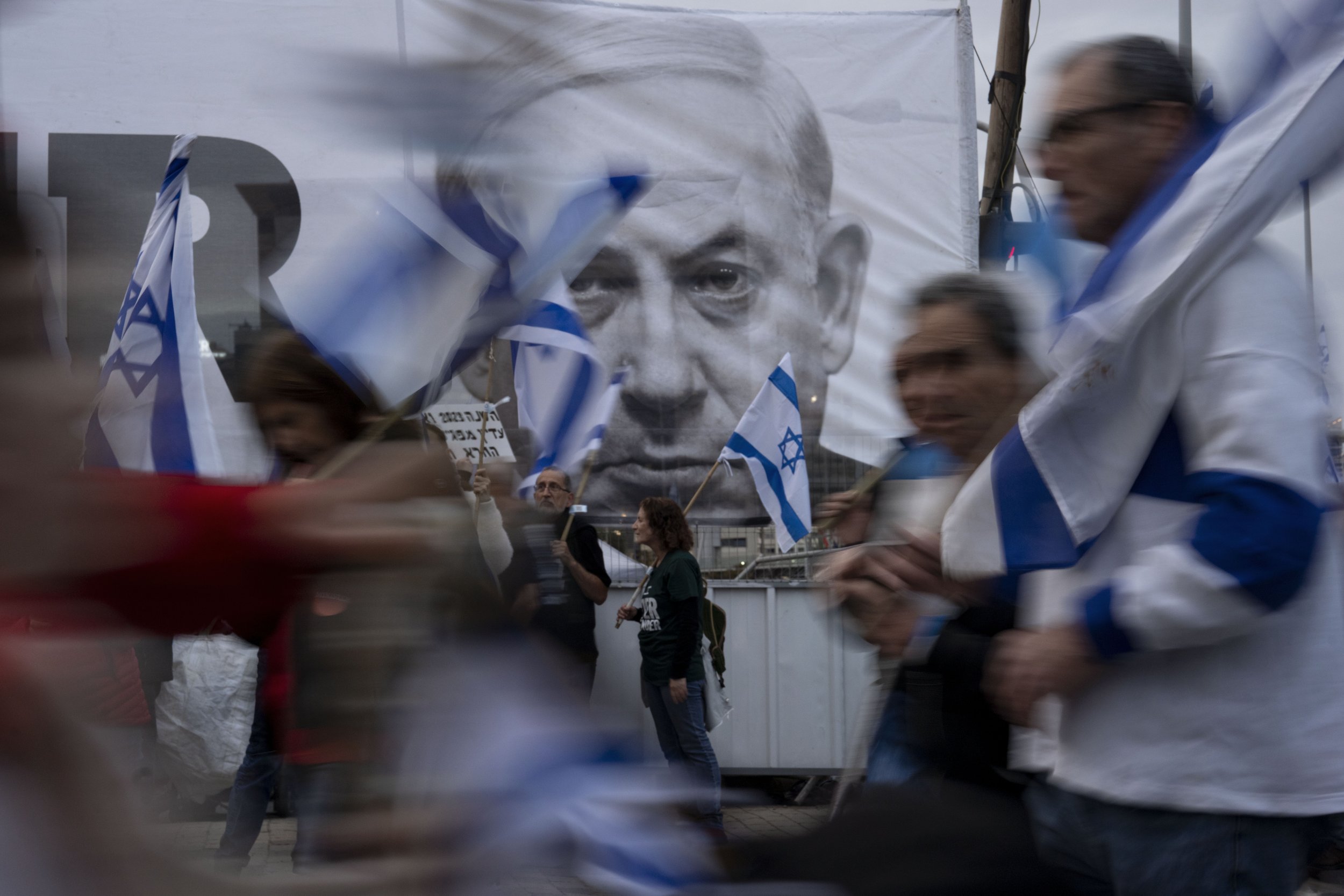  Demonstrators walk with Israel's national flags next to a banner showing Prime Minister Benjamin Netanyahu during a protest against plans by his government to overhaul the judicial system in Tel Aviv, Israel, on March 25, 2023. (AP Photo/Oded Balilt