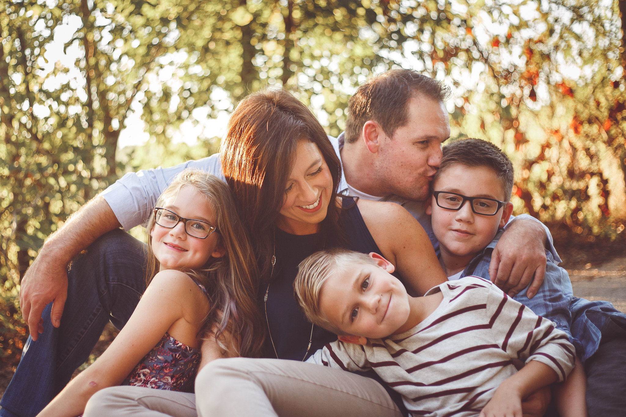 A family of 5 cuddles together and is all smiles with the trees and evening sunshine over their shoulders in a family photograph by L+R Photography