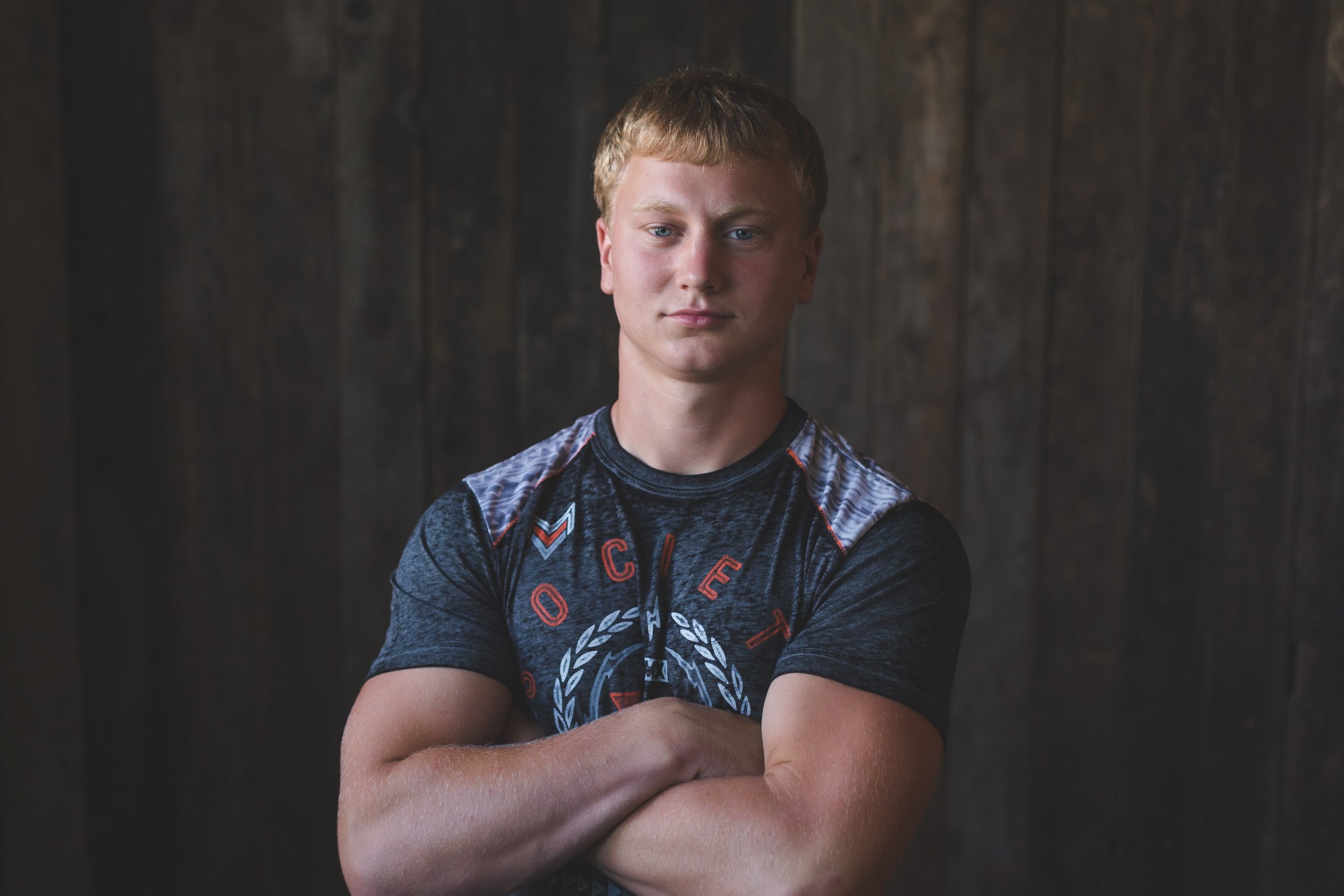 A young senior guy poses for the camera in a t-shirt and in front of a wood background in this senior photography session by L+R Photography - Tulsa , Oklahoma