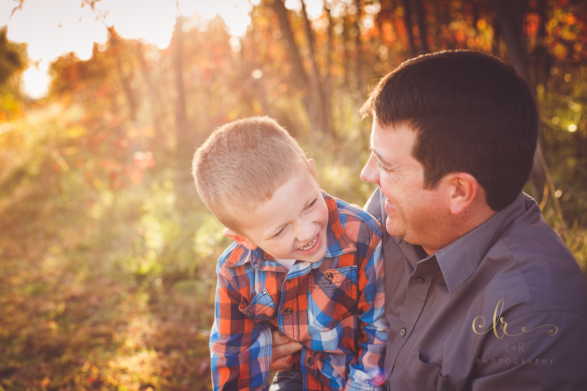 A young boy wearing read and blue flannel is right at home in his father's arms with the beautiful autumn leaves and sunset behind them - Tulsa Oklahoma Family Photography