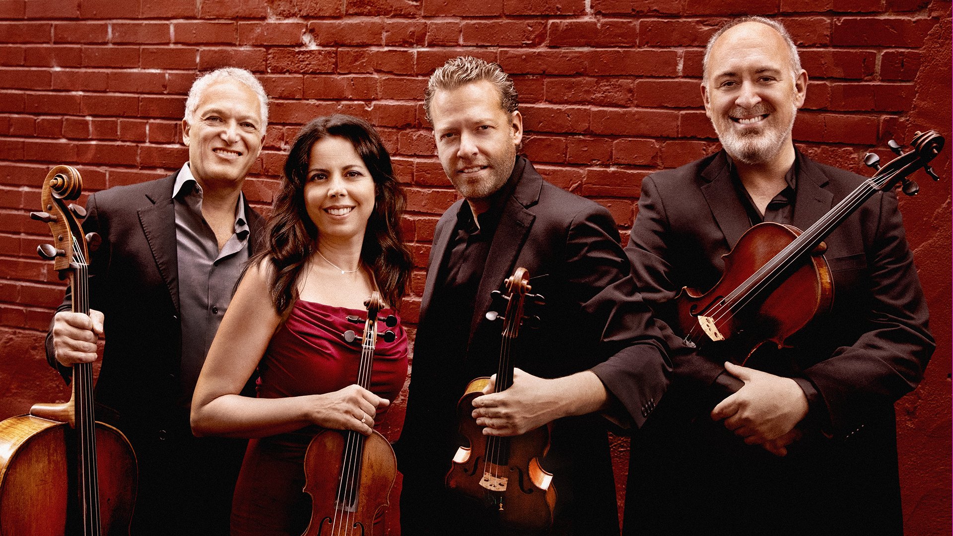 Four musicians stand against a brick wall holding various string instruments