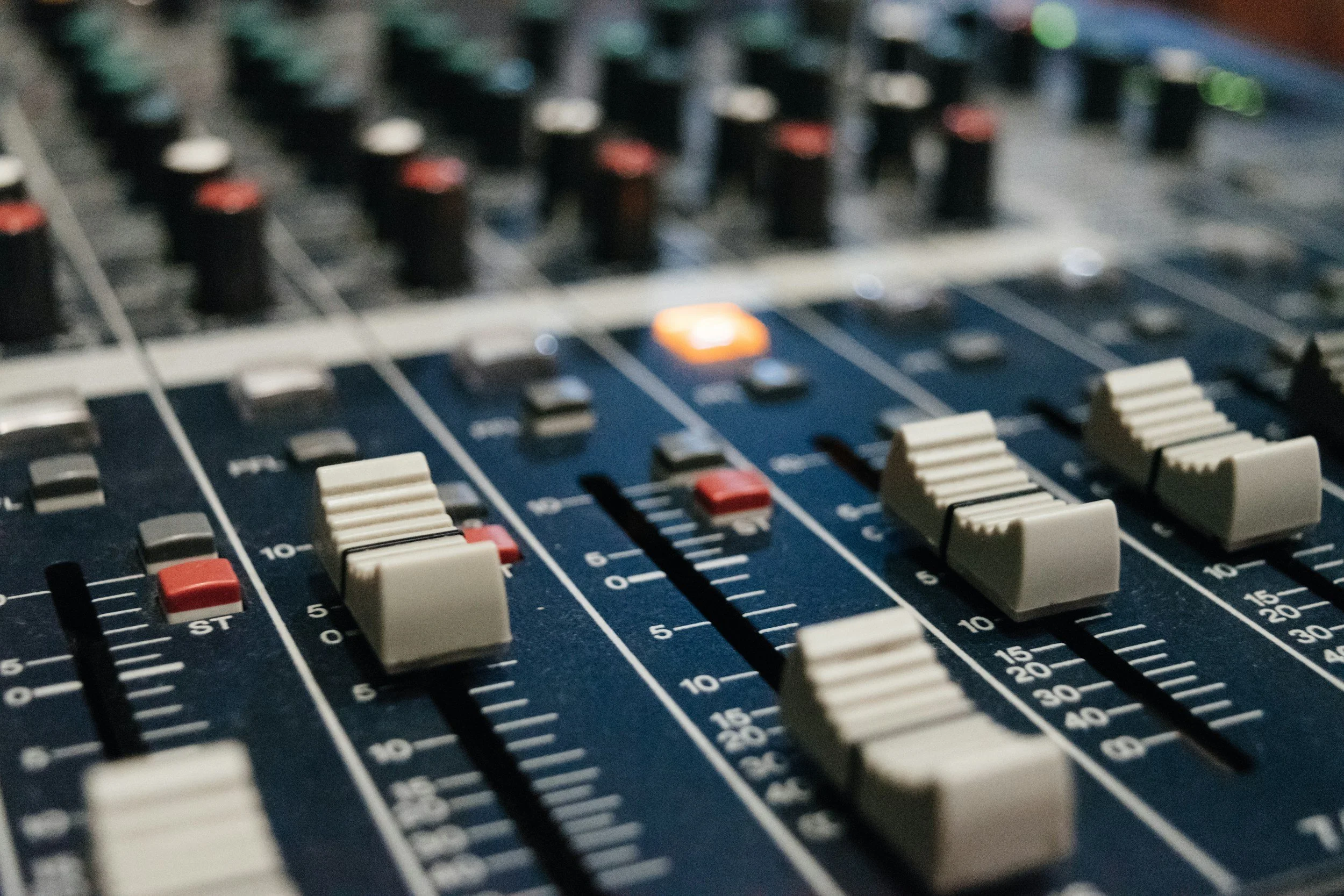 A close-up of an audio mixing console with various faders, knobs, and buttons, some illuminated, used for sound control.