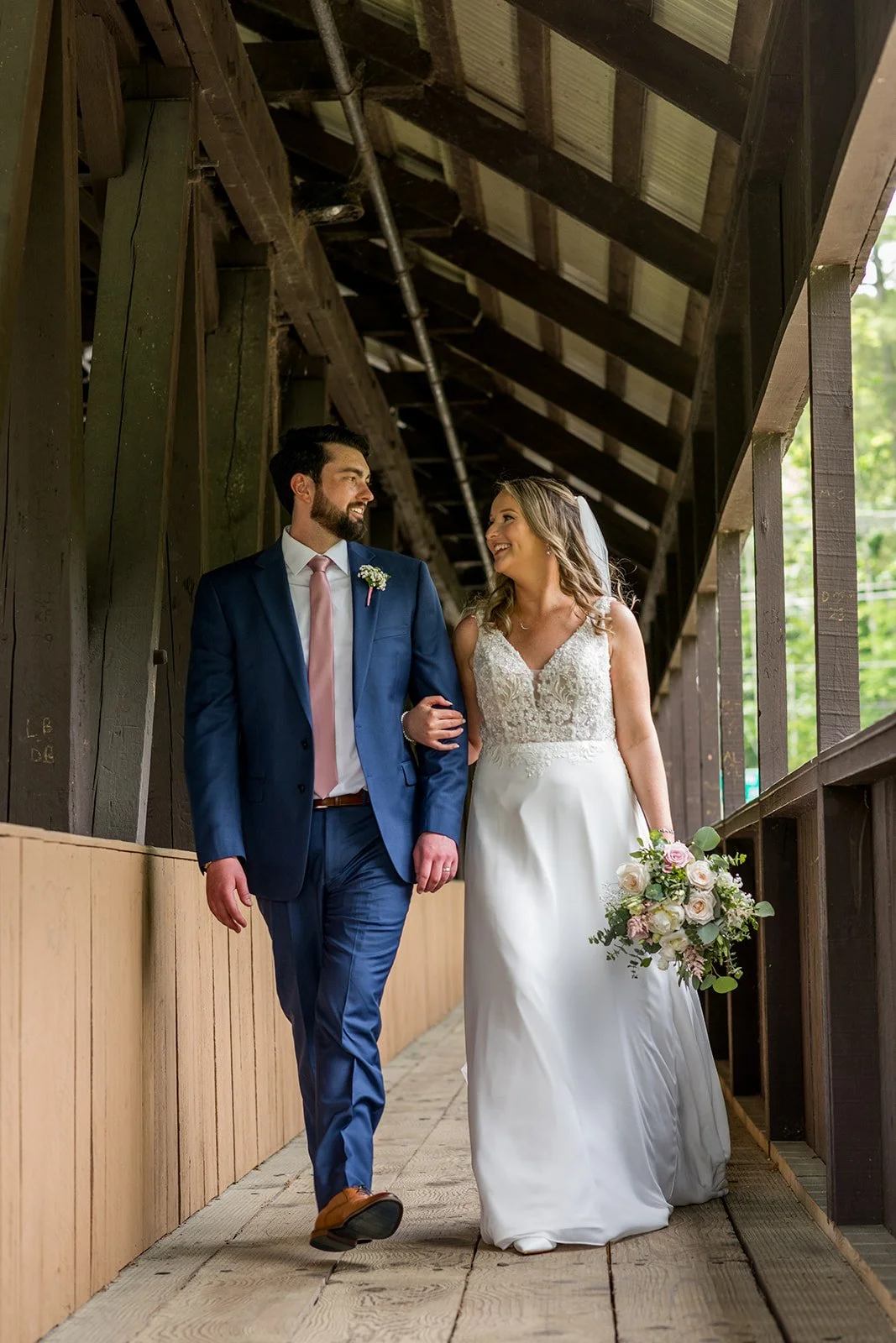 Bride and groom walking together on a wooden walkway during their wedding, smiling at each other, with the bride holding a bouquet of flowers.