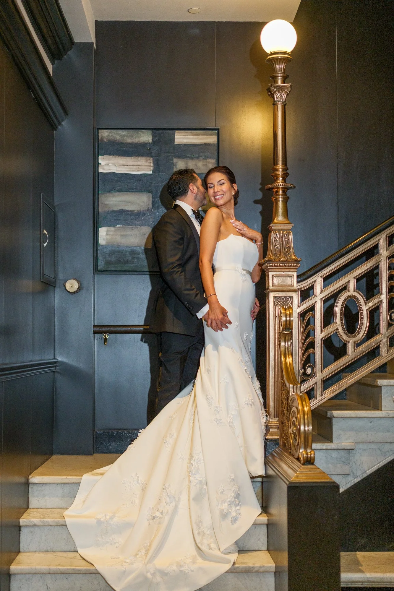 Groom whispering to smiling bride on the elegant gold-railed staircase inside the XV Beacon Hotel Boston