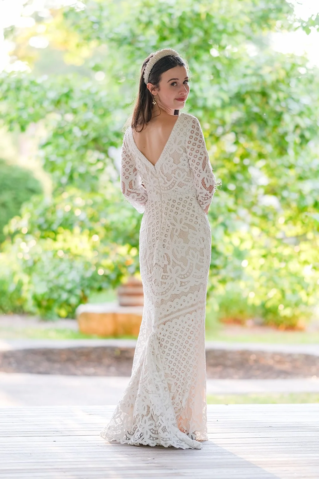 A woman in a white lace dress with long sleeves, standing outdoors with greenery in the background.