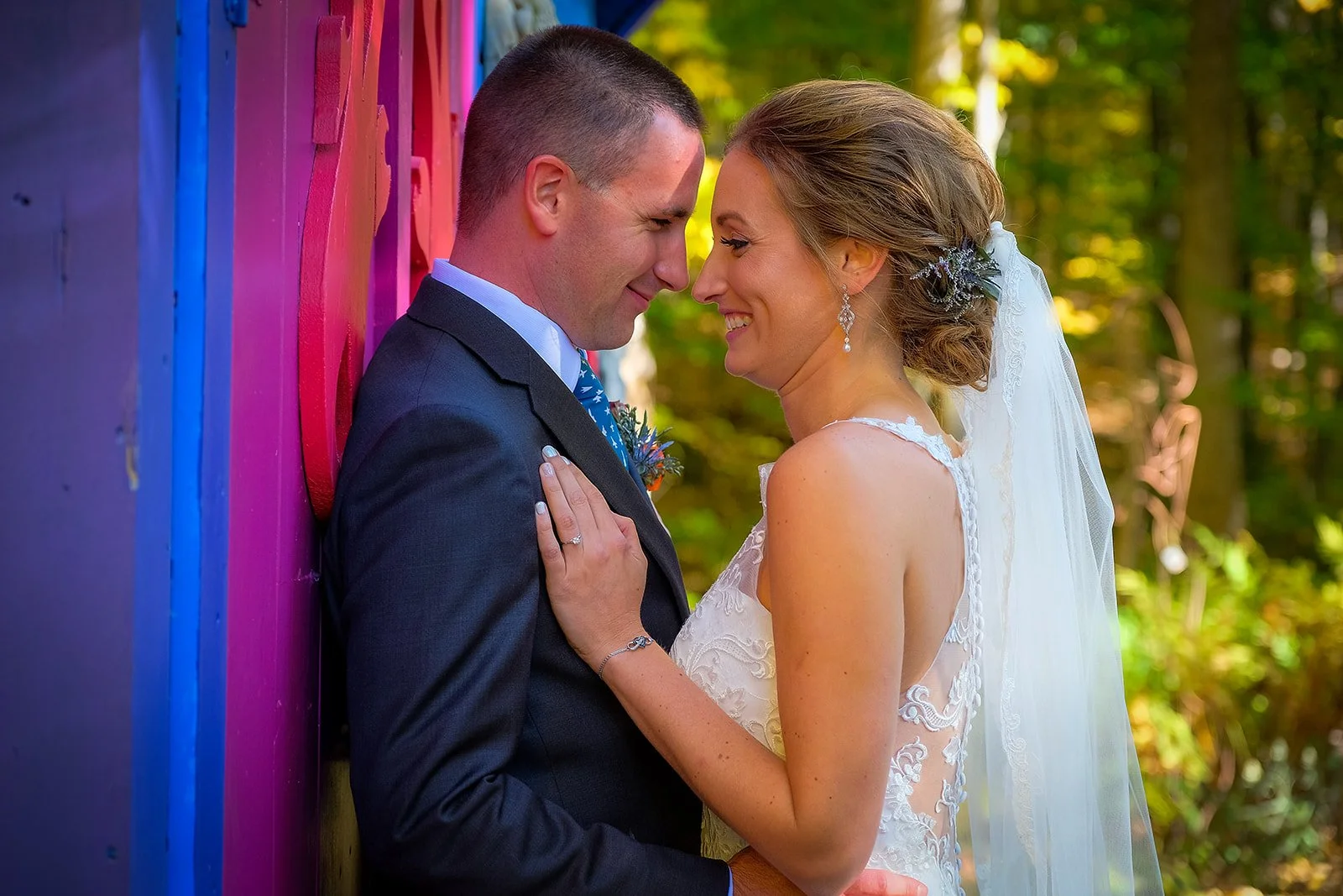 A bride and groom standing close together outdoors, leaning their foreheads against each other with smiling faces, in a colorful wooded setting.