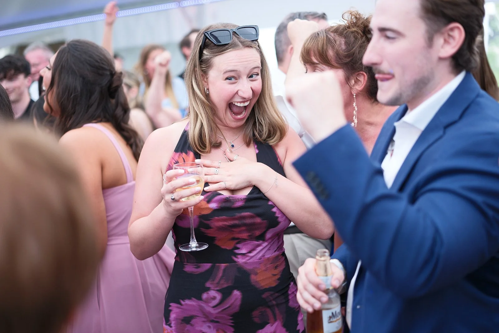 A woman with blonde hair, wearing a floral dress and sunglasses on her head, is smiling and holding a drink at a celebration or party. She appears to be surprised or excited, with her hand on her chest, in a crowd of people dressed in formal attire.