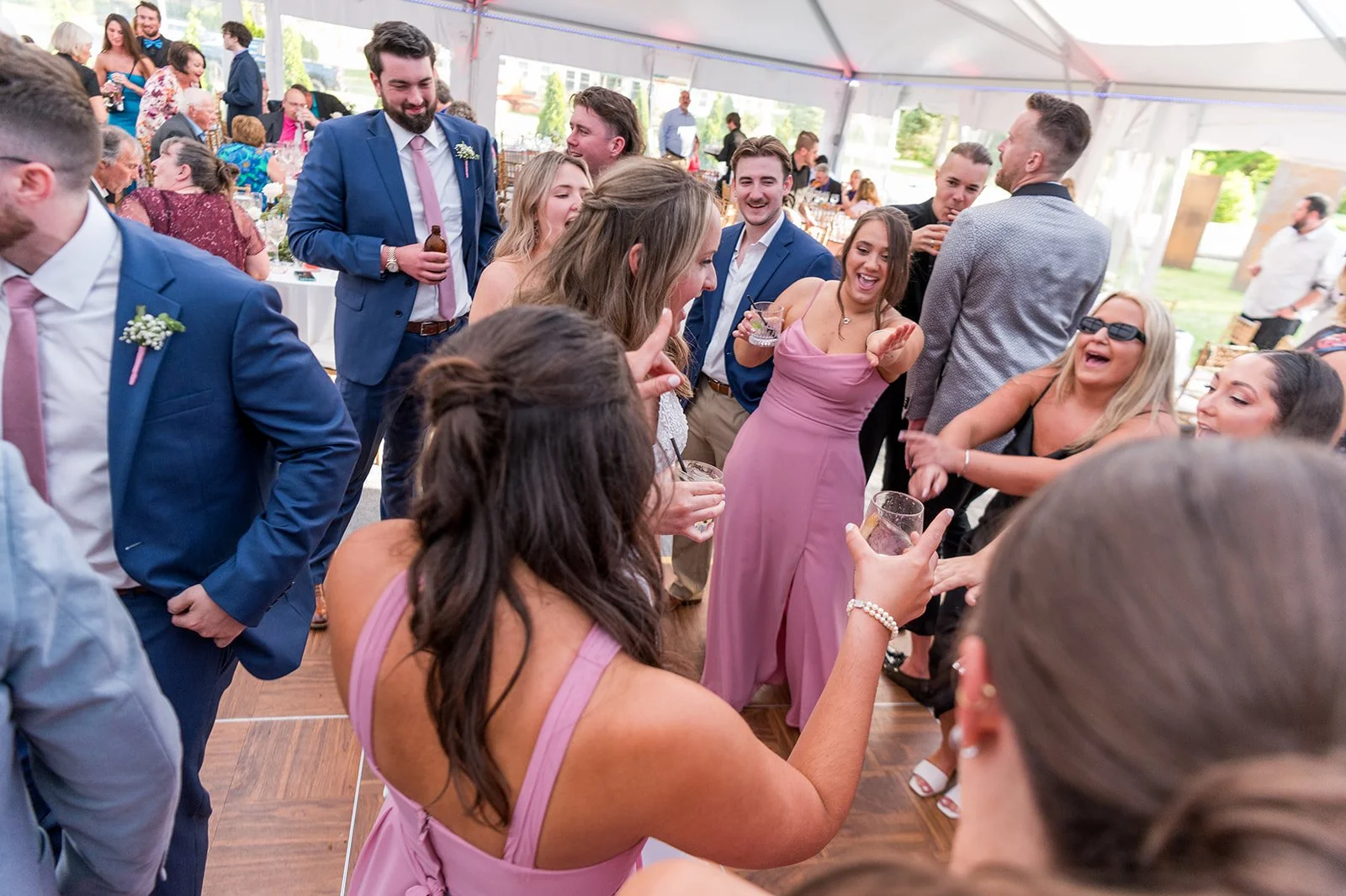 People celebrating and dancing at a wedding reception inside a large tent, dressed in formal attire, with tables, chairs, and decorations visible in the background.