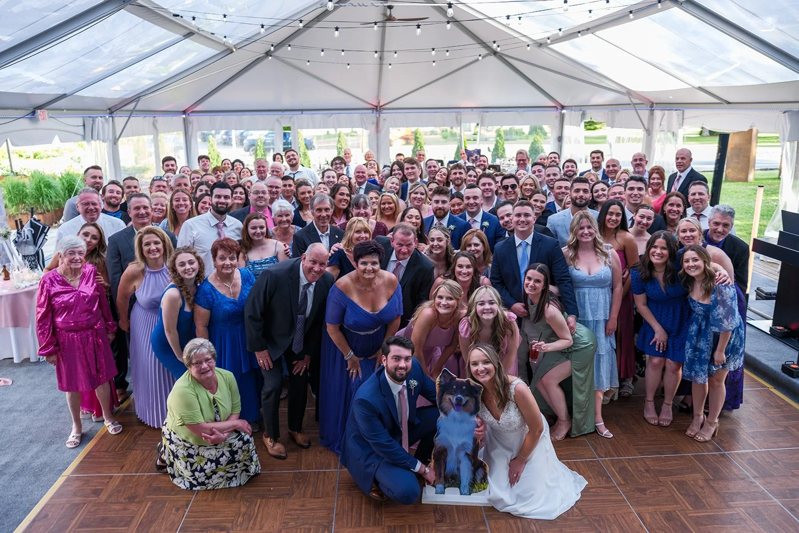 A large group of people at a wedding reception inside a white tent, with a bride and groom at the front, kneeling with a dog. The guests are smiling and dressed in formal attire, with greenery and outdoor scenery visible through the tent windows.