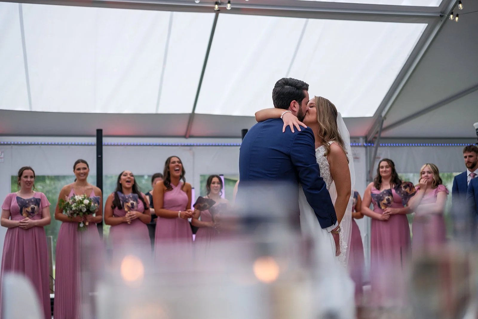 A couple kissing at their wedding reception under a tent, with bridesmaids and groomsmen in the background smiling and laughing.