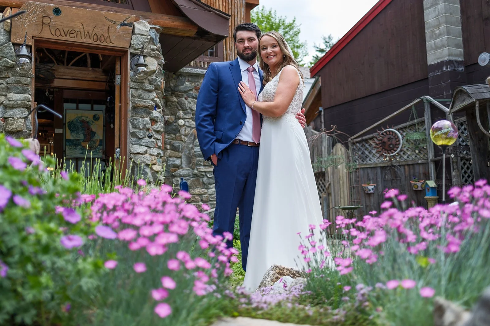 A newlywed couple standing in a garden filled with pink flowers, smiling for the camera, with a stone and wood building labeled "Ravenwood" in the background.