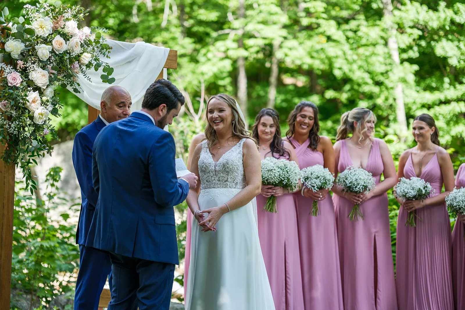 A wedding ceremony taking place outdoors with the bride and groom exchanging vows, surrounded by bridesmaids in pink dresses holding white bouquets, and an officiant and groomsmen present. The scene is set in a green, wooded area with a floral arch