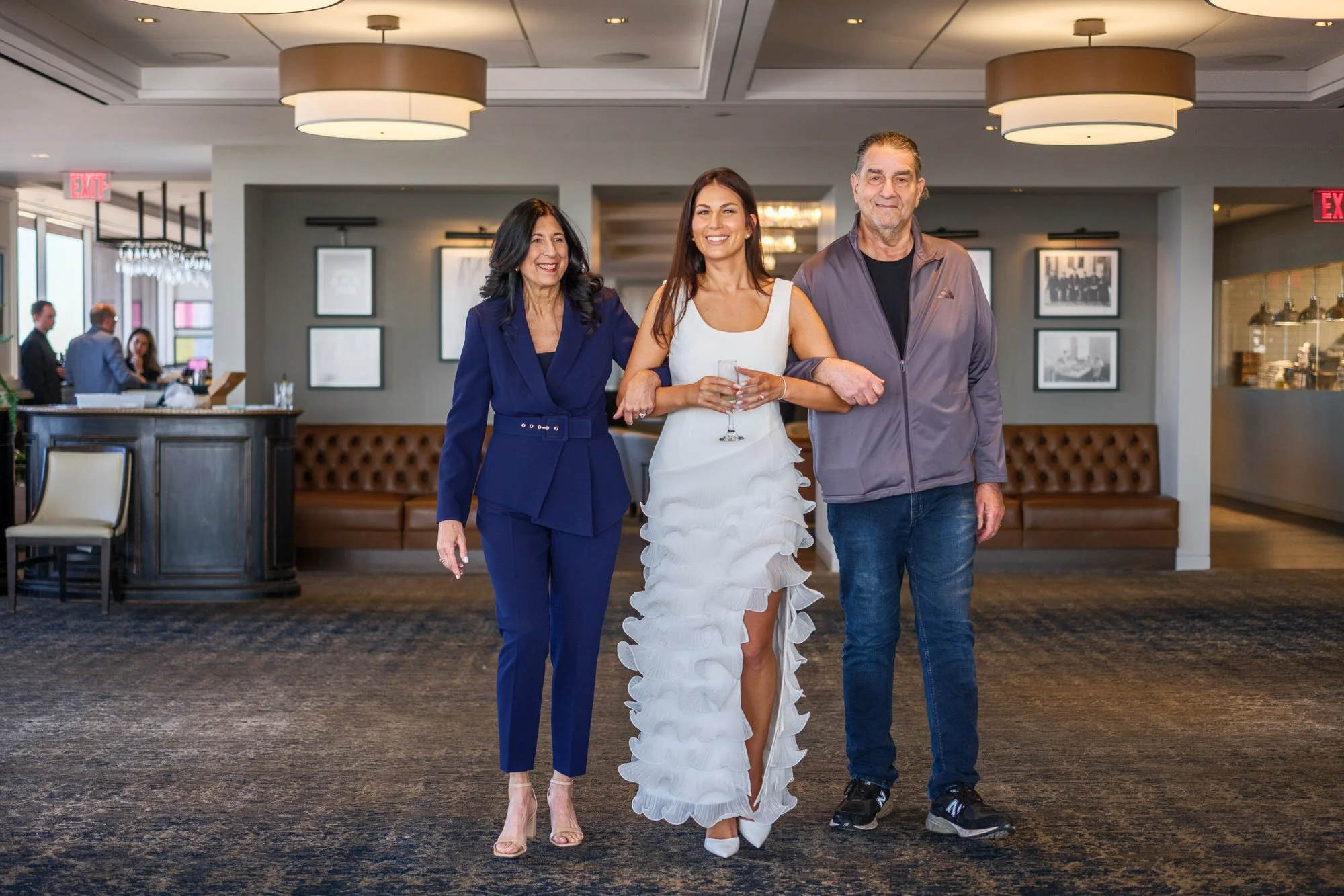 Lauren in her white ruffle dress holding a champagne flute, standing with her mother and father in the UMass Club event space just before her rehearsal dinner